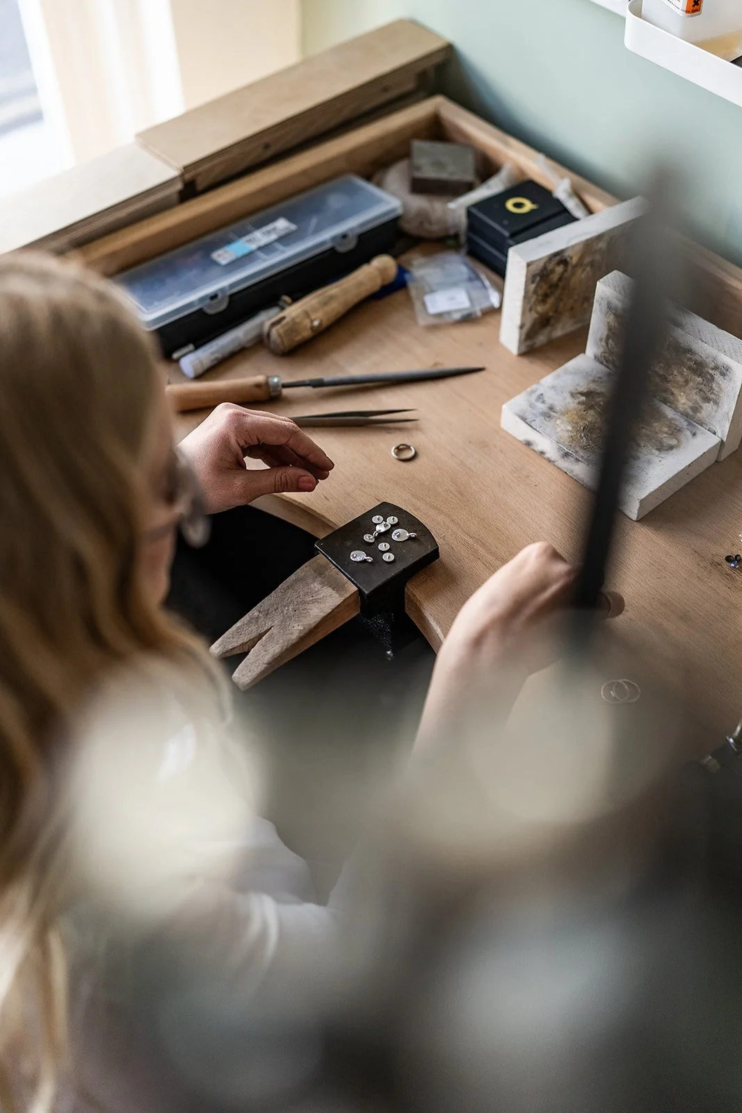 A woman working on jewelry at a jewelry workbench with tools, stones, and jewelry pieces.