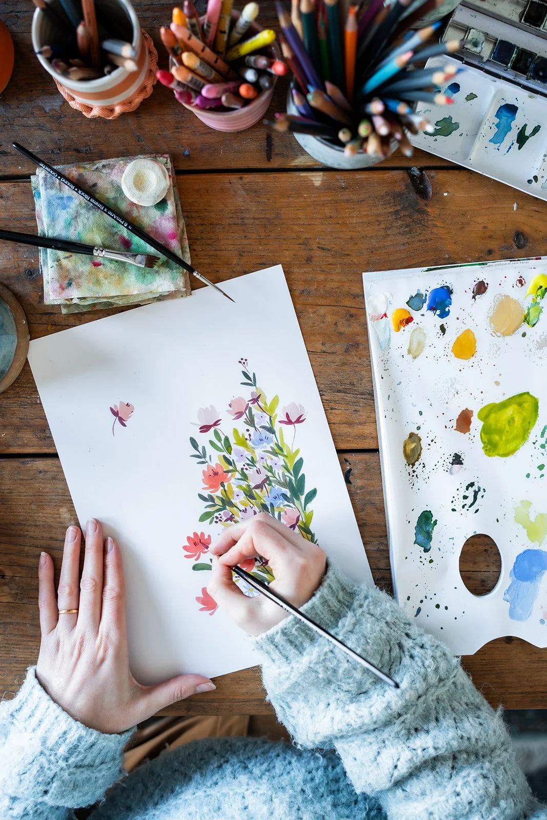 Person painting a floral watercolor artwork on paper, surrounded by paint supplies and brushes on a wooden table.