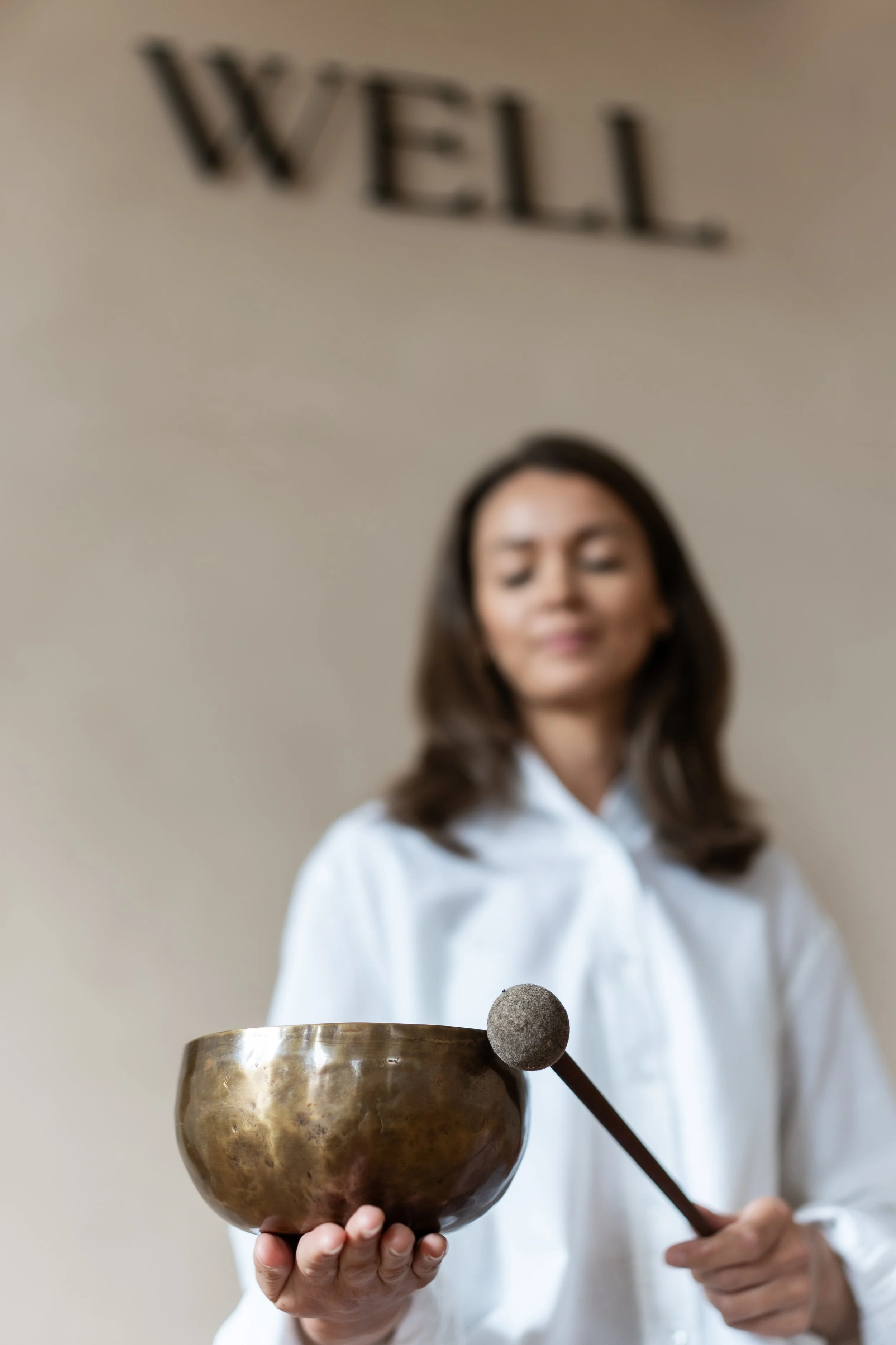 A woman holding a Tibetan singing bowl and mallet, with the "WELL" sign in the background.