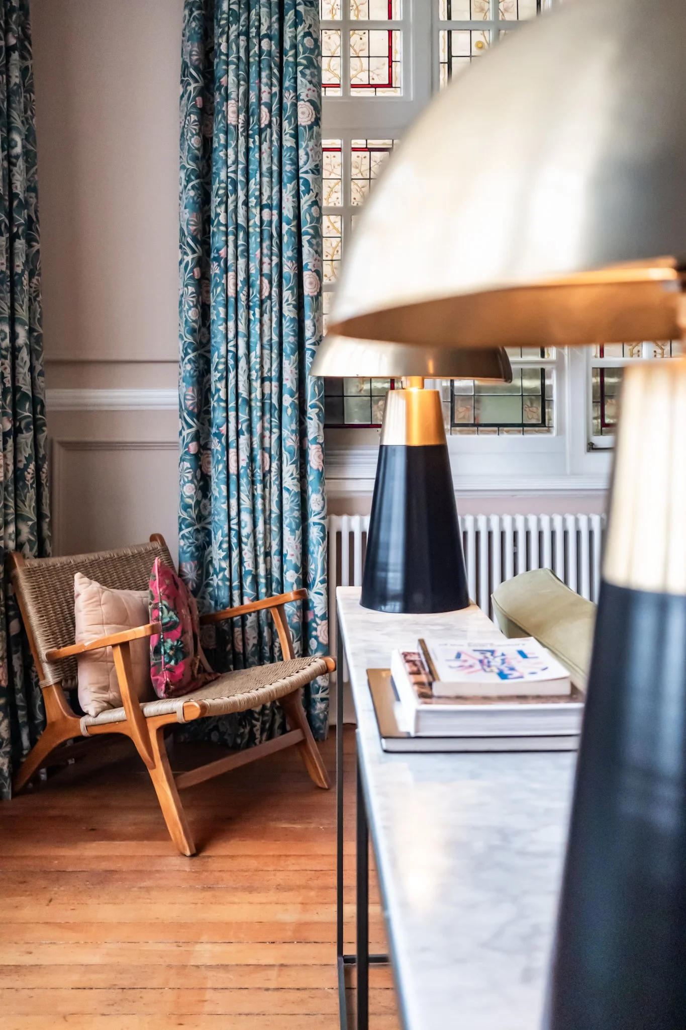 Interior of a room with blue floral curtains, a wooden chair with a pink floral pillow, a marble-topped table with lamps and books, a window with stained glass, and wooden flooring.