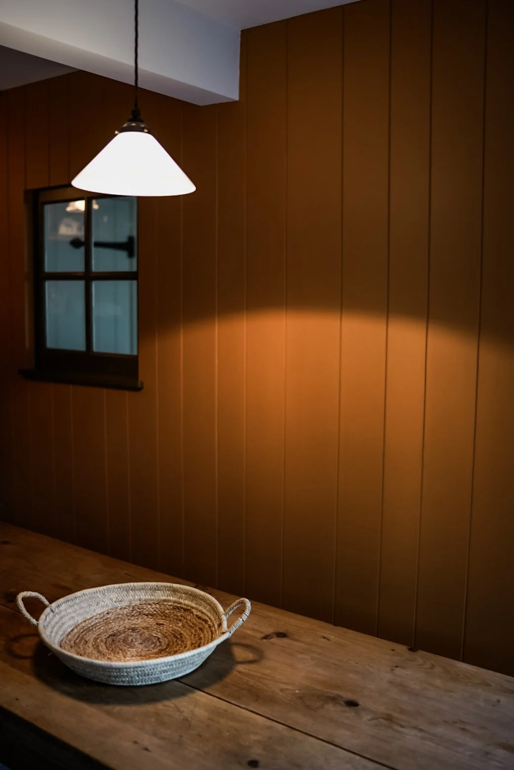 A wooden table with a woven basket, a brown wooden panel wall, a small window, and a hanging white lamp.
