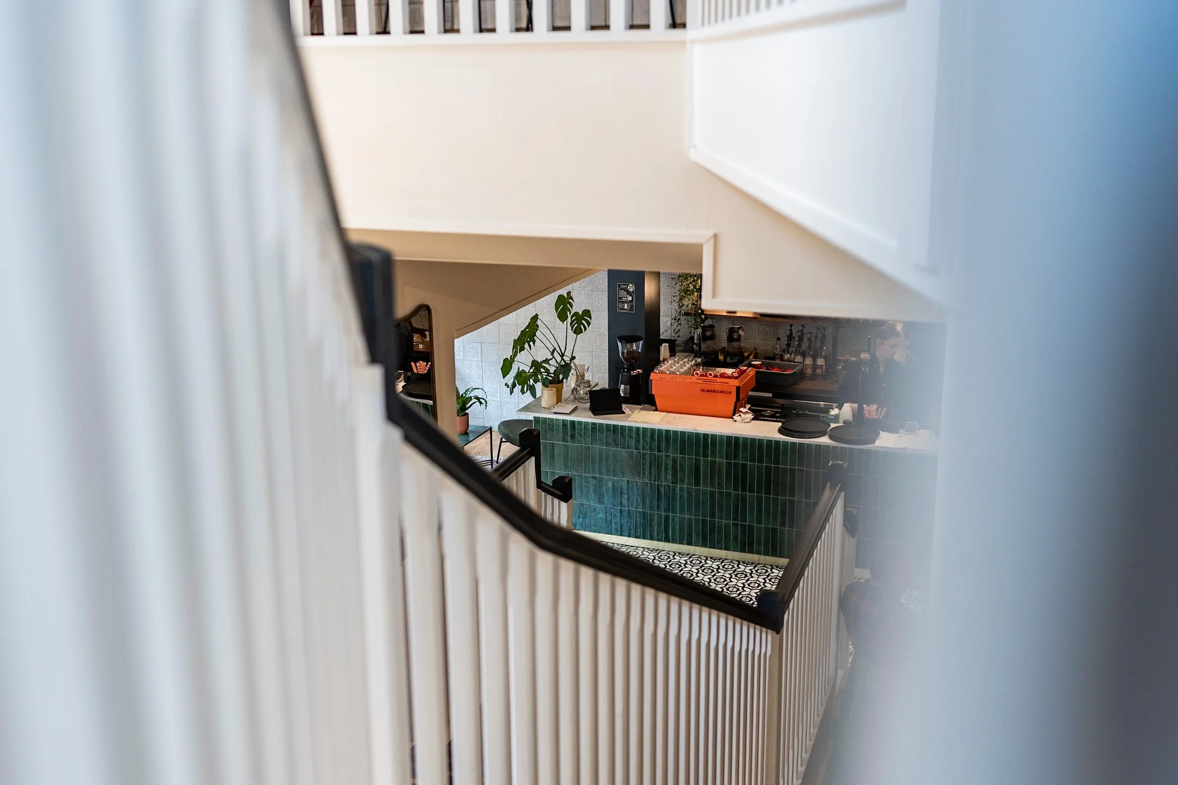 View from a staircase looking down into a cafe area with green tiled counter, potted plants, coffee machine, and baristas working behind the counter.