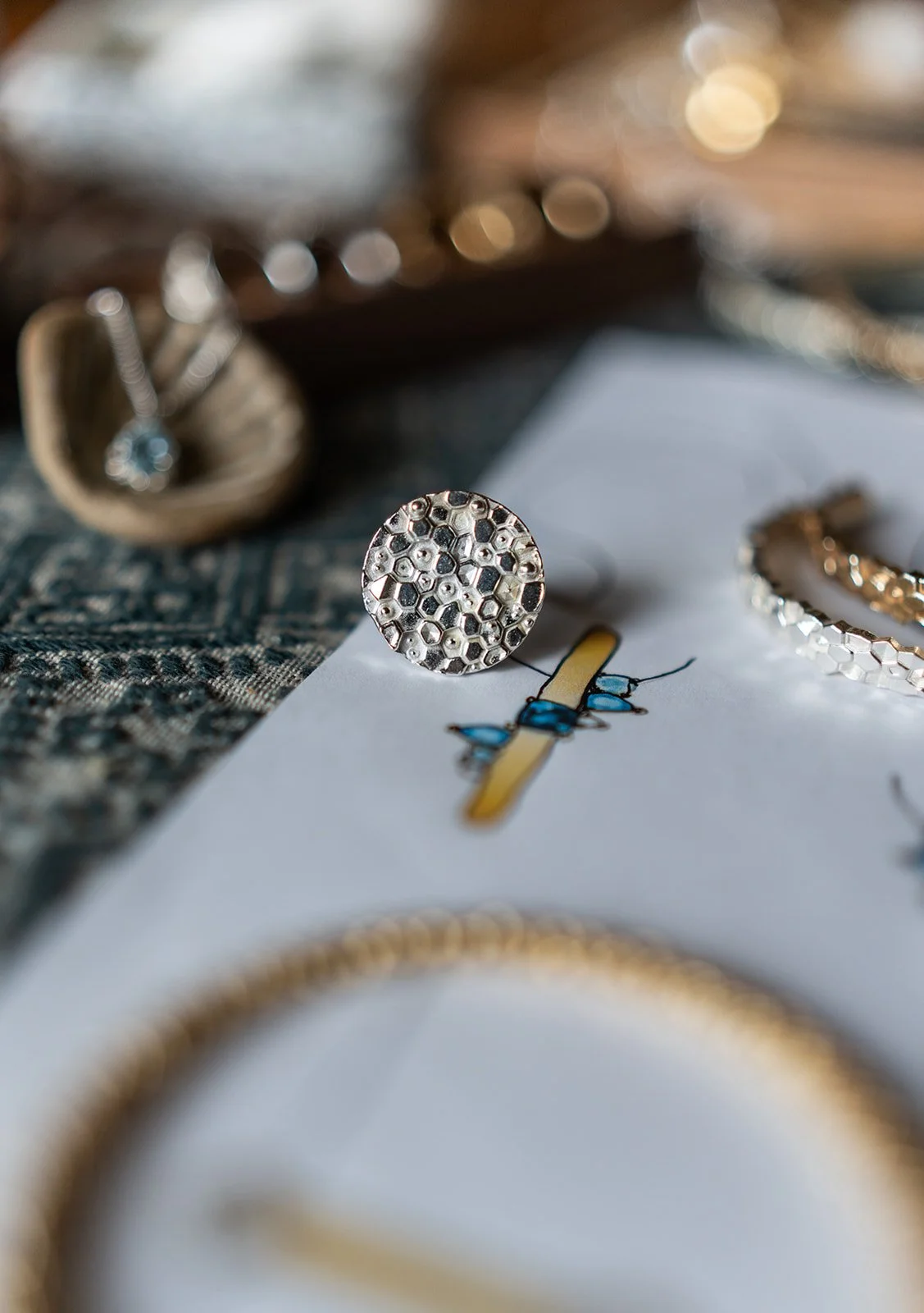 Close-up of silver jewelry, including a round honeycomb-patterned ring, placed on a drawing of a yellow bug with blue wings, with blurred background of more jewelry and a gold chain.
