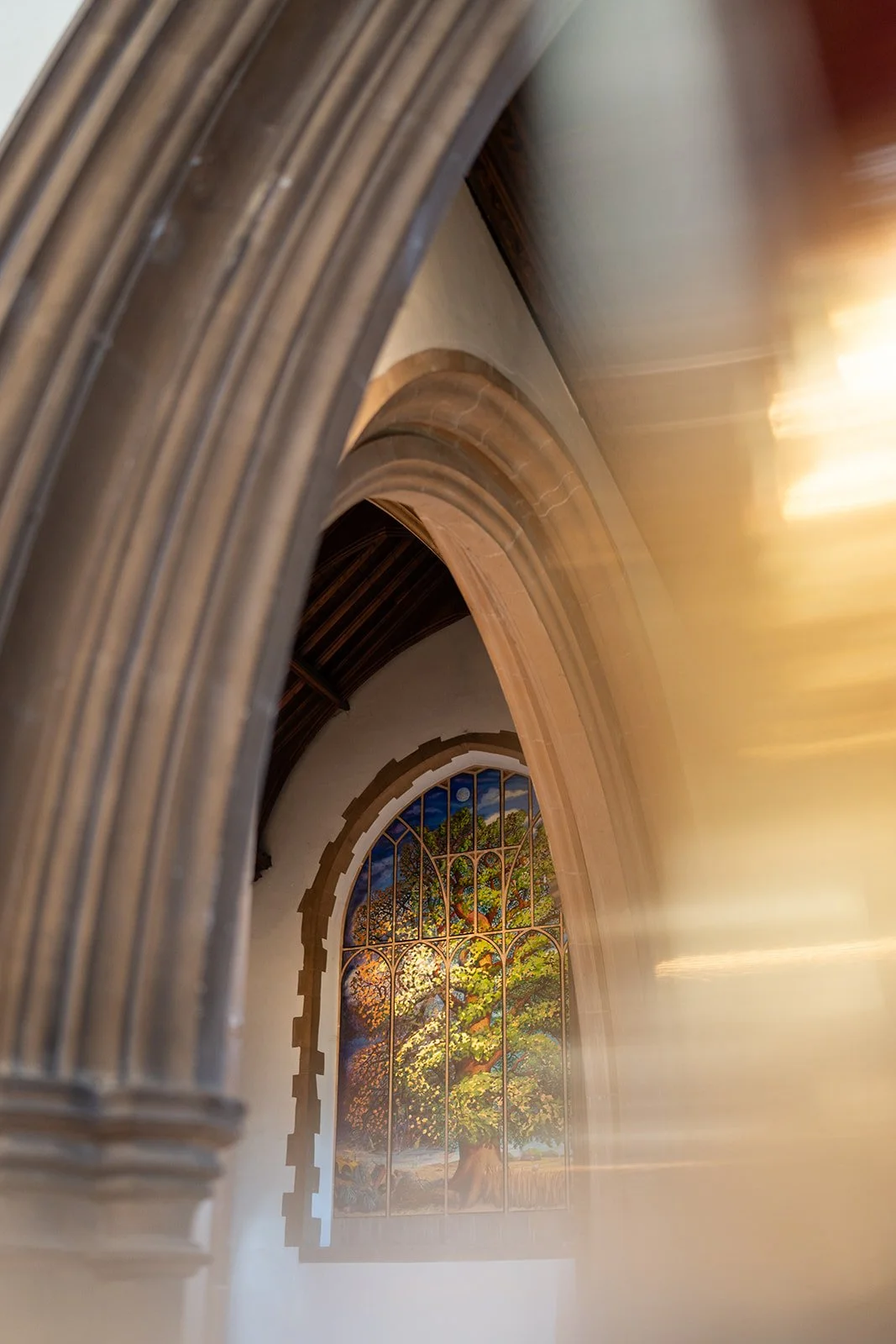 Interior view of a church or cathedral, focusing on a stained glass window depicting a large tree with colorful leaves, surrounded by stone arches and wooden ceiling.