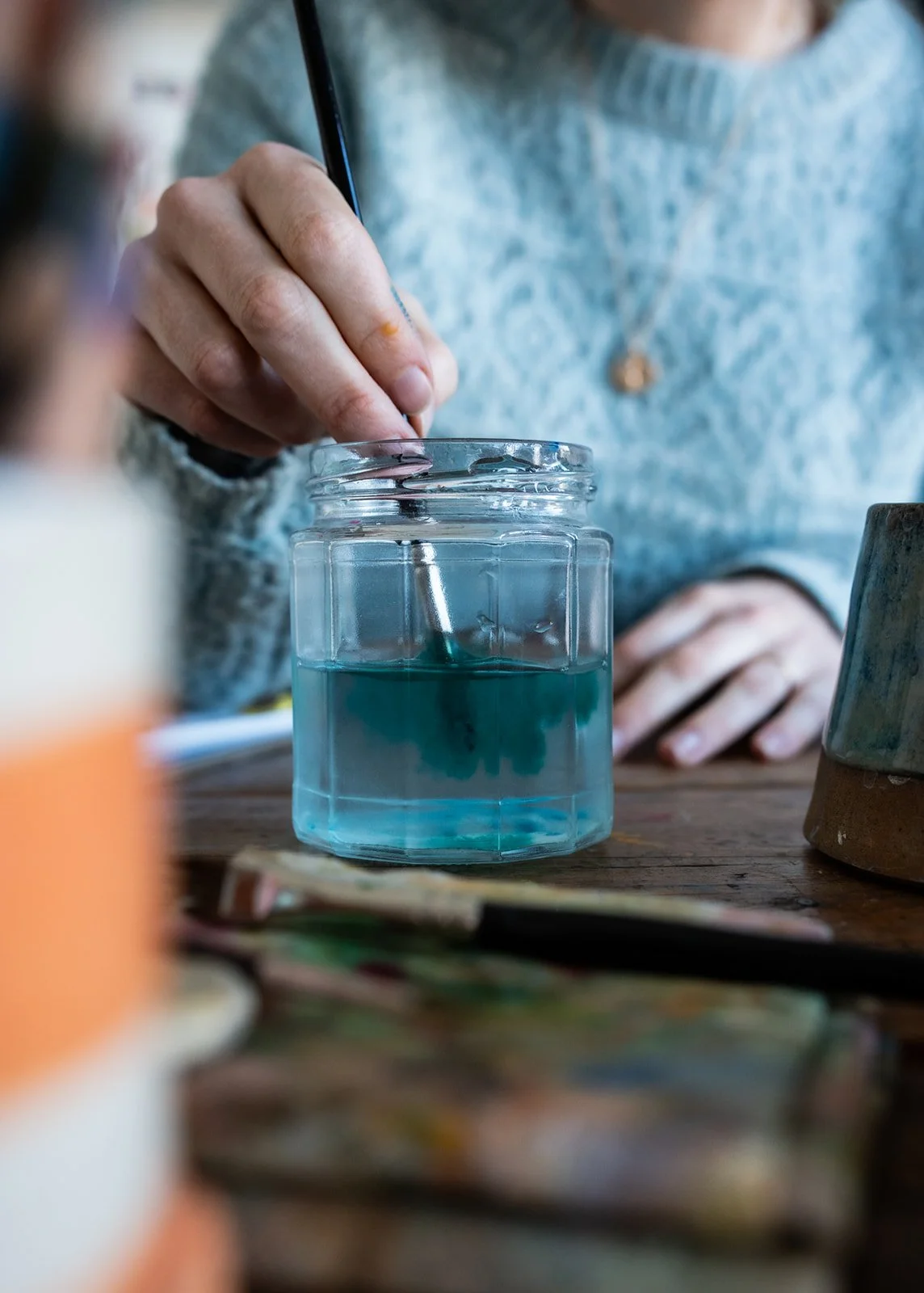 Person using a pen to write or draw in a glass jar filled with blue liquid, on a wooden table.