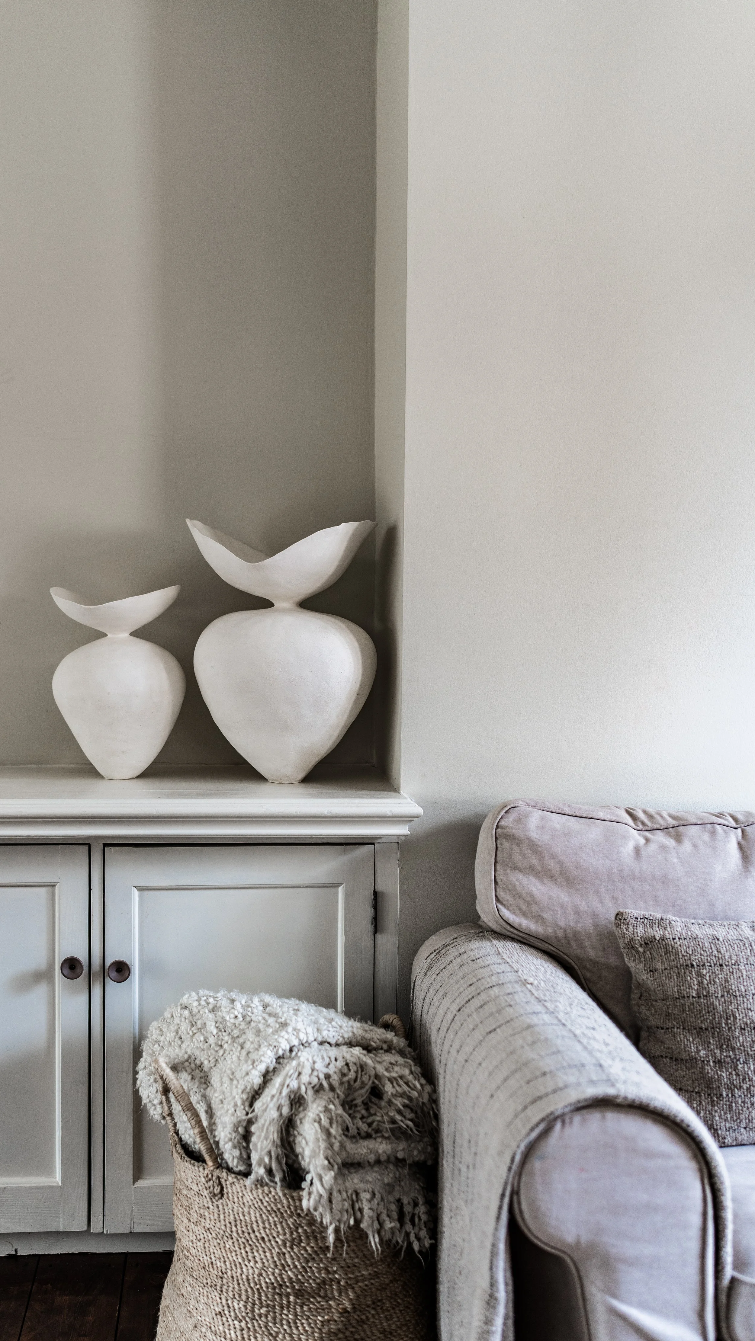 Decorative white ceramic vases on a white cabinet in a neutral-toned living room with a soft beige sofa and a woven basket with a throw blanket.