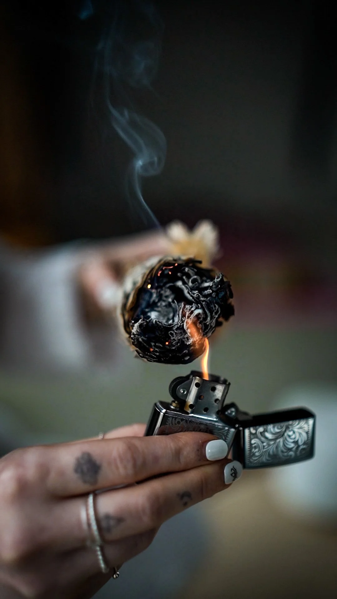 Close-up of a hand holding a lighter, with a burning piece of charcoal or wood. Smoke is rising from the fire.