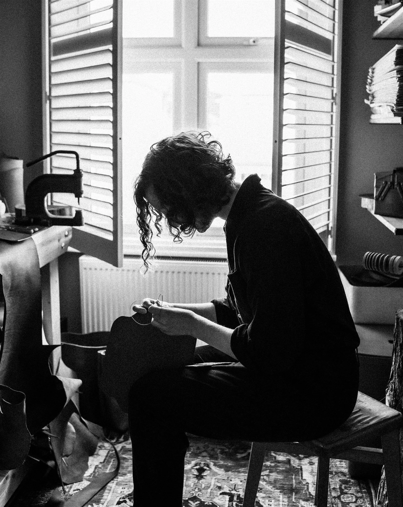 A person sitting on a chair in a room with a window, working on a sewing project, with shelves of fabric and a sewing machine nearby.
