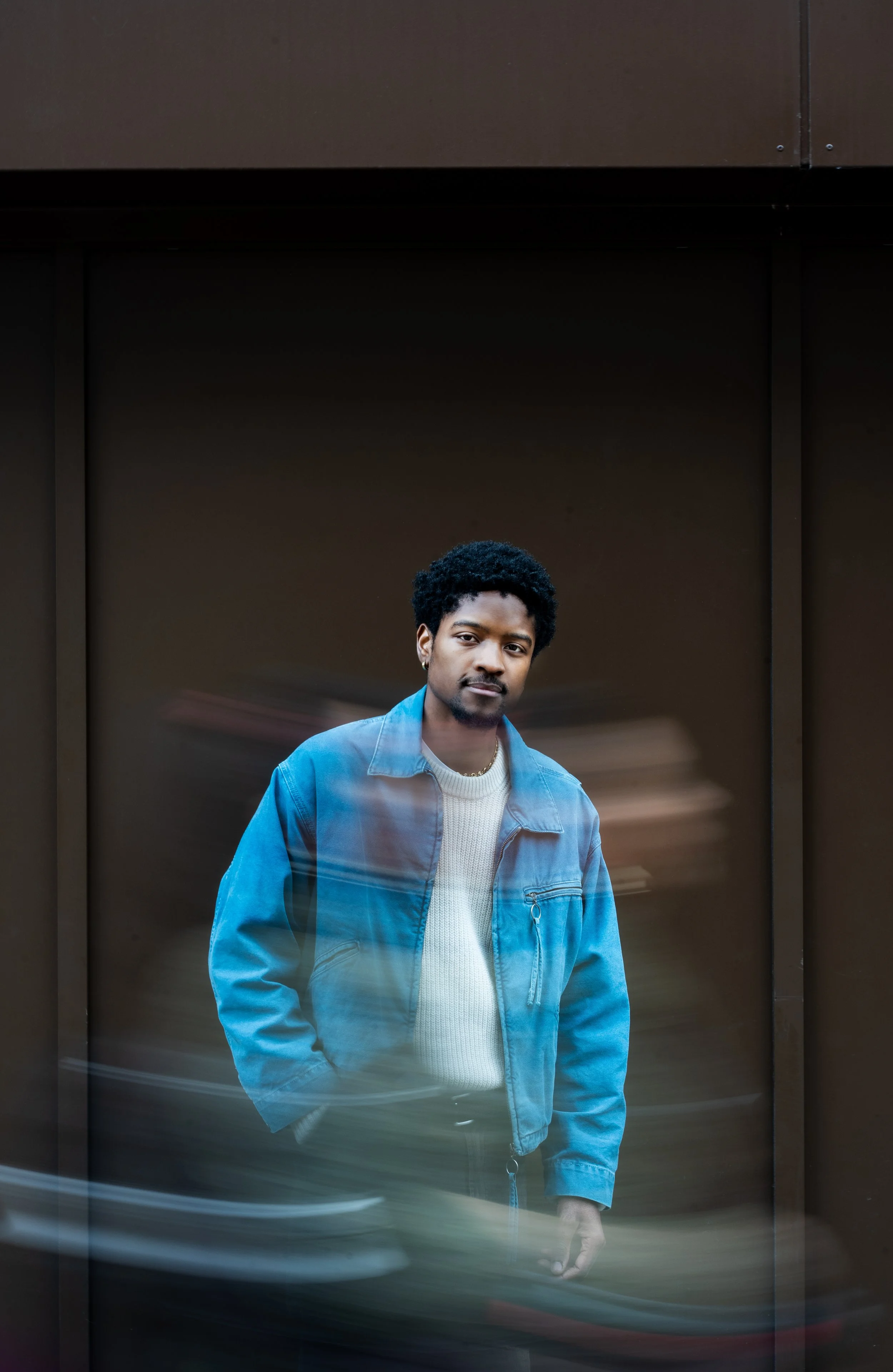 A young man with dark curly hair and a mustache, wearing a white sweater and a blue jacket, stands in front of a dark, modern exterior wall. There are blurred streaks of light across the image, possibly from passing cars.
