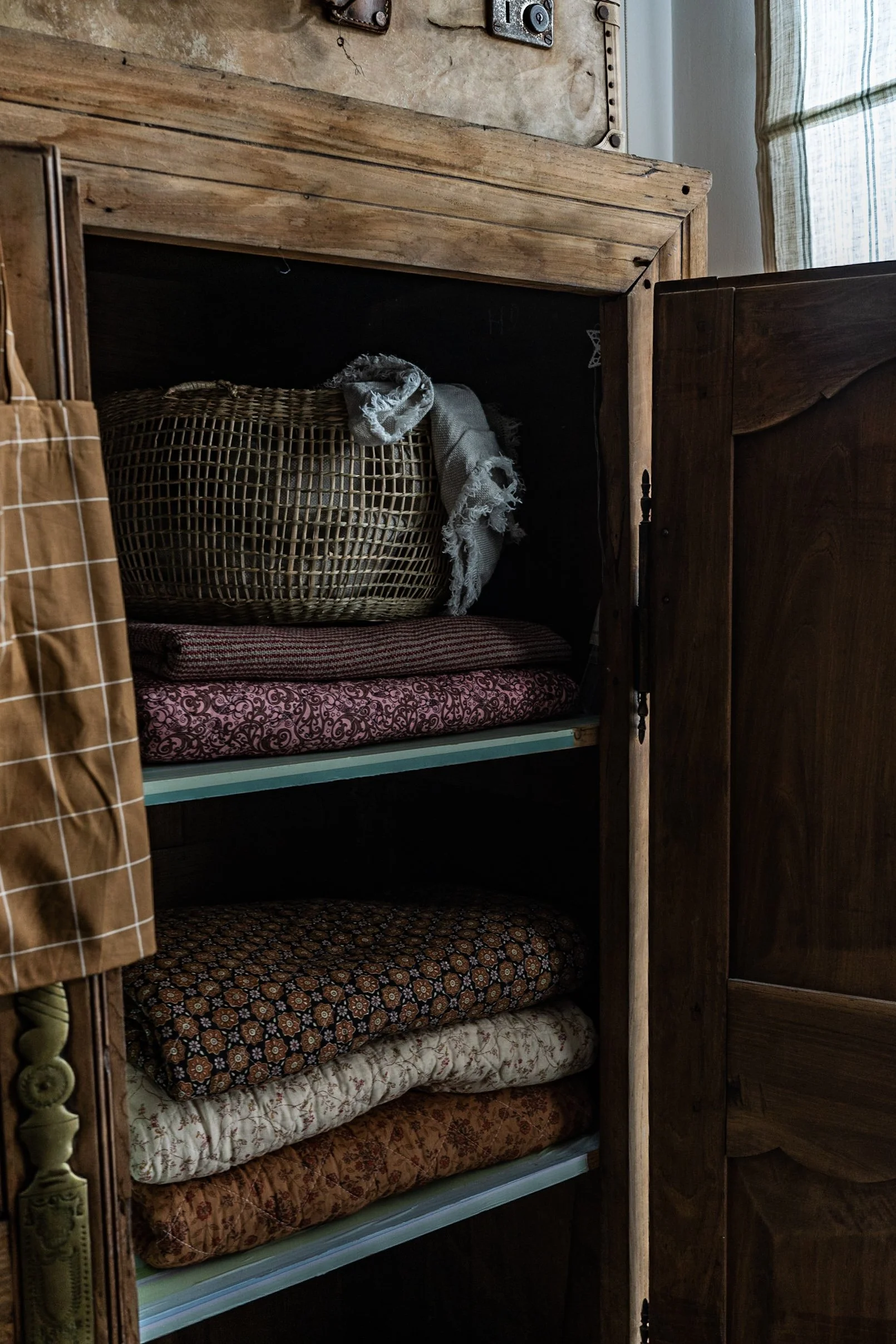 An open wooden cabinet filled with folded blankets and a woven basket on top, all against a rustic wall with a switchboard.