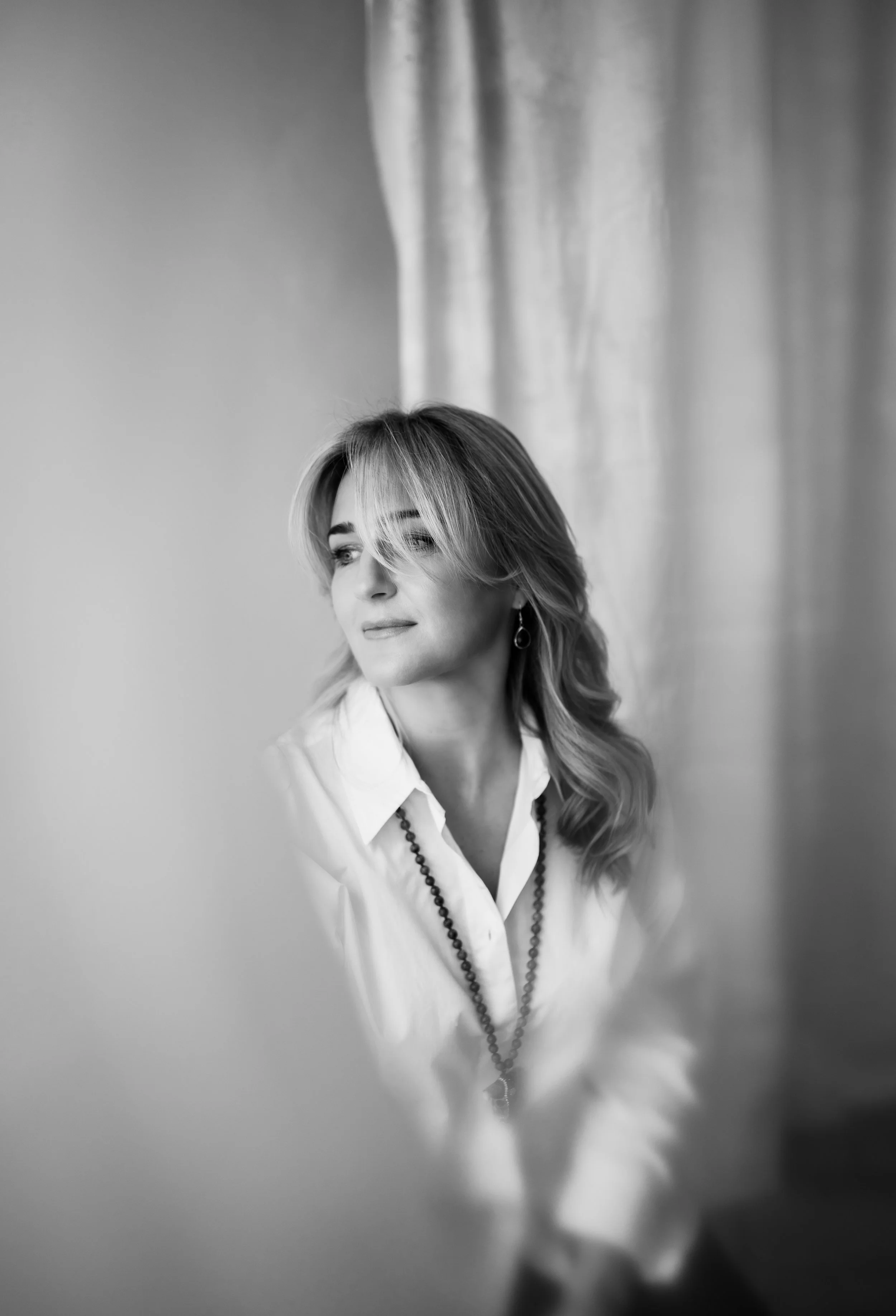 Black and white portrait of a woman with shoulder-length hair, looking to the side, wearing a white shirt, earrings, and a long necklace, sitting in front of a curtain.