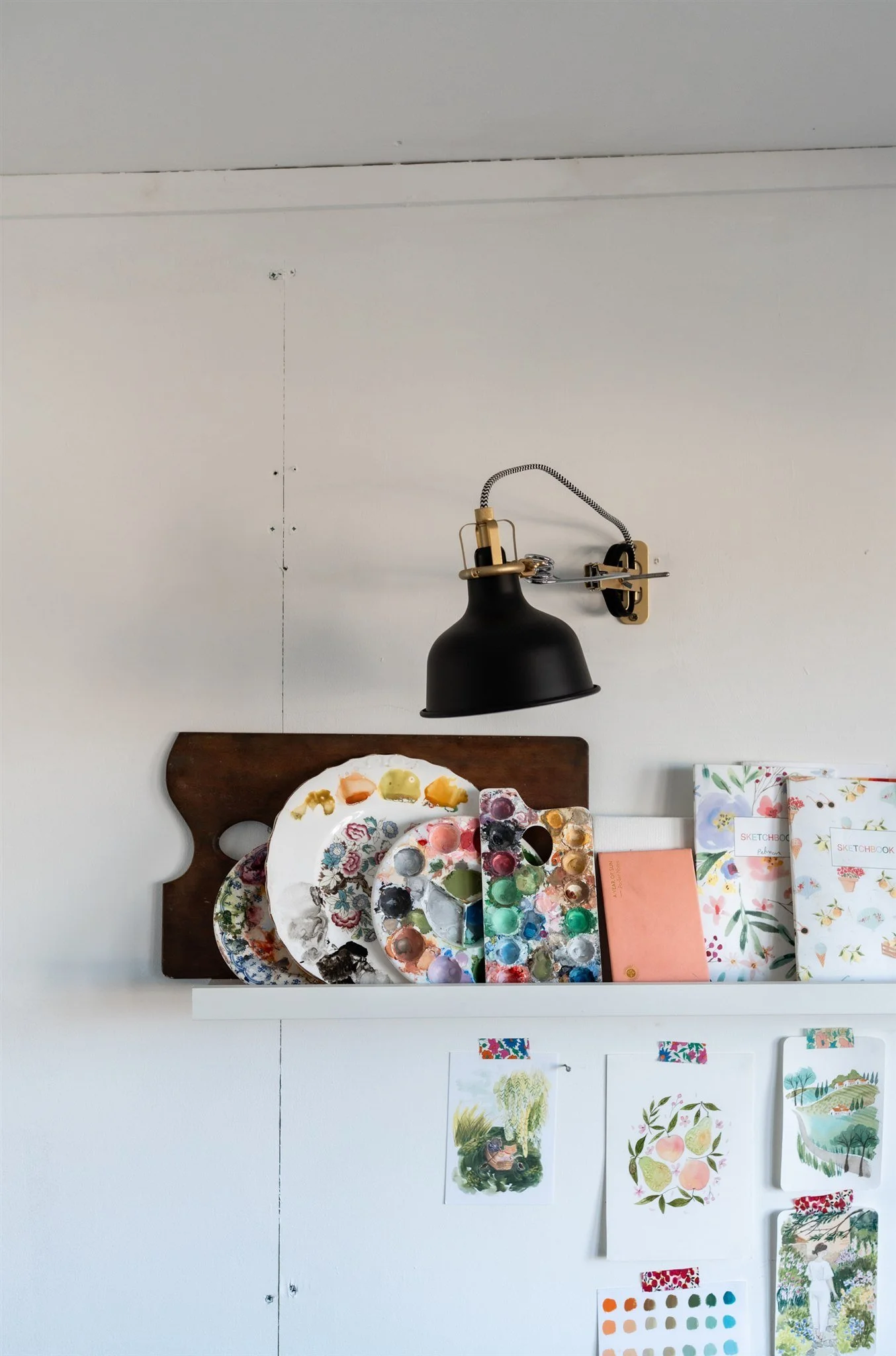 An art setup with a black wall-mounted lamp above a shelf displaying painted palettes, notebooks, and sketchbooks, with colorful artwork and prints taped to the white wall below.