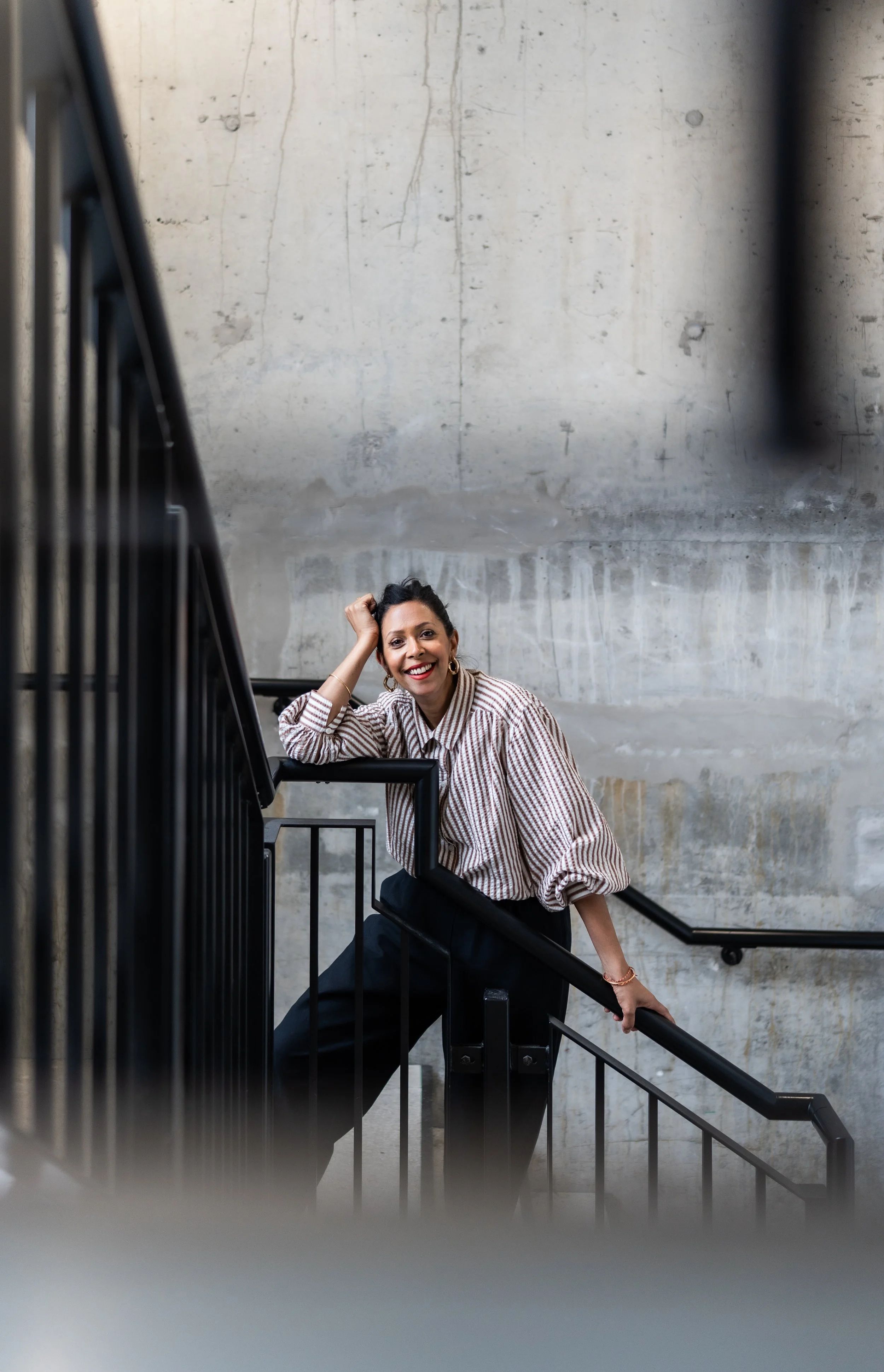 A woman smiling at the camera, leaning on a staircase railing, with a concrete wall background.