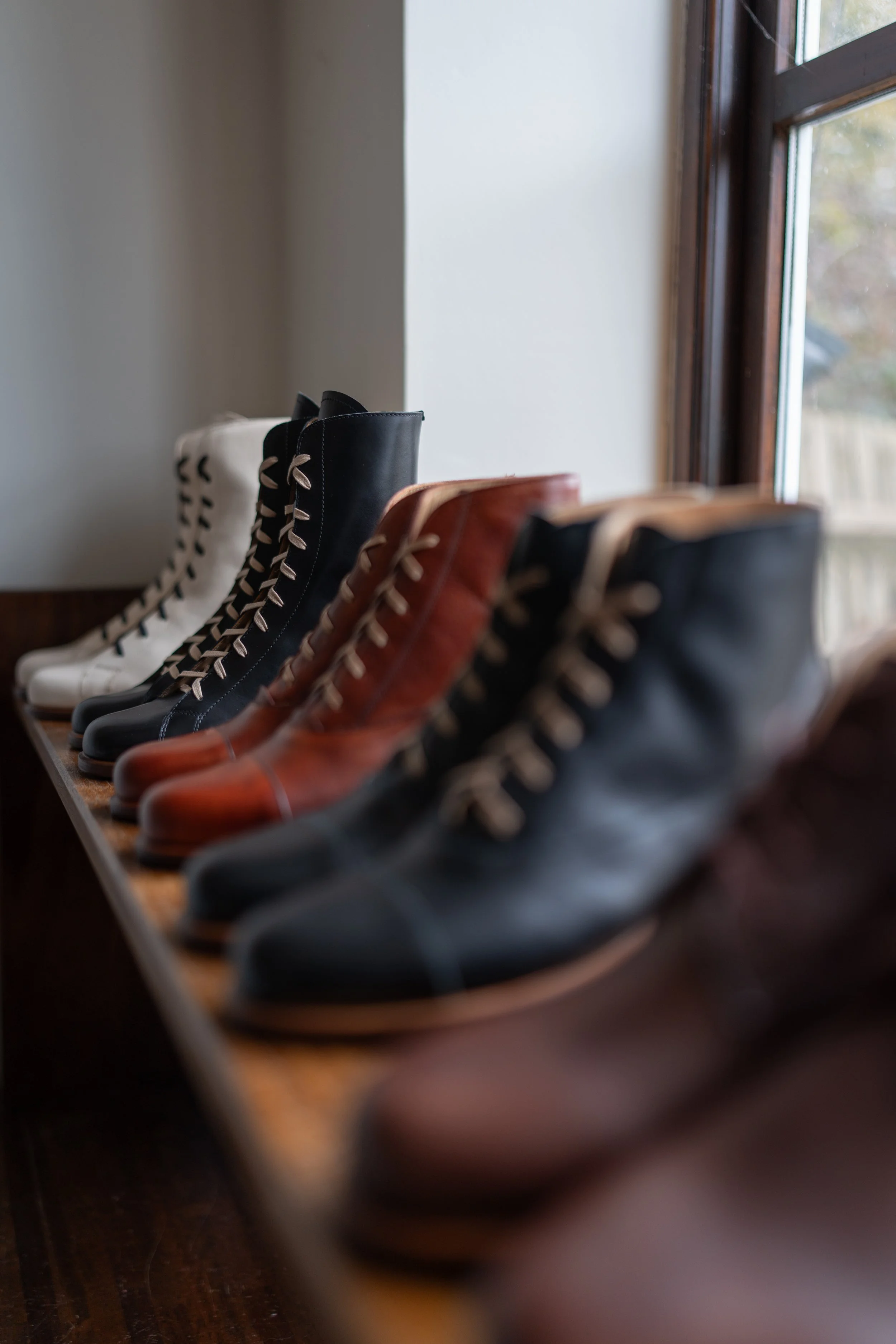 A row of vintage leather boots in black, brown, and white, lined up on a wooden surface by a window.