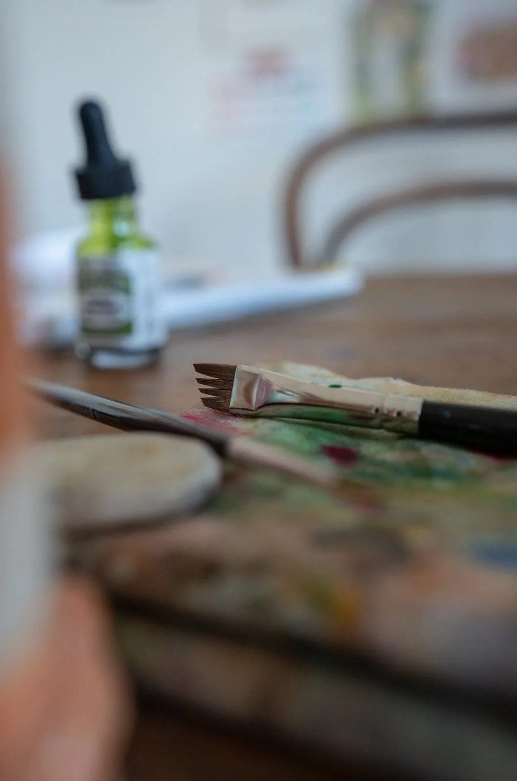 Close-up of paintbrushes and an artist's palette on a table with a blurred background of a bottle of paint and a whiteboard.