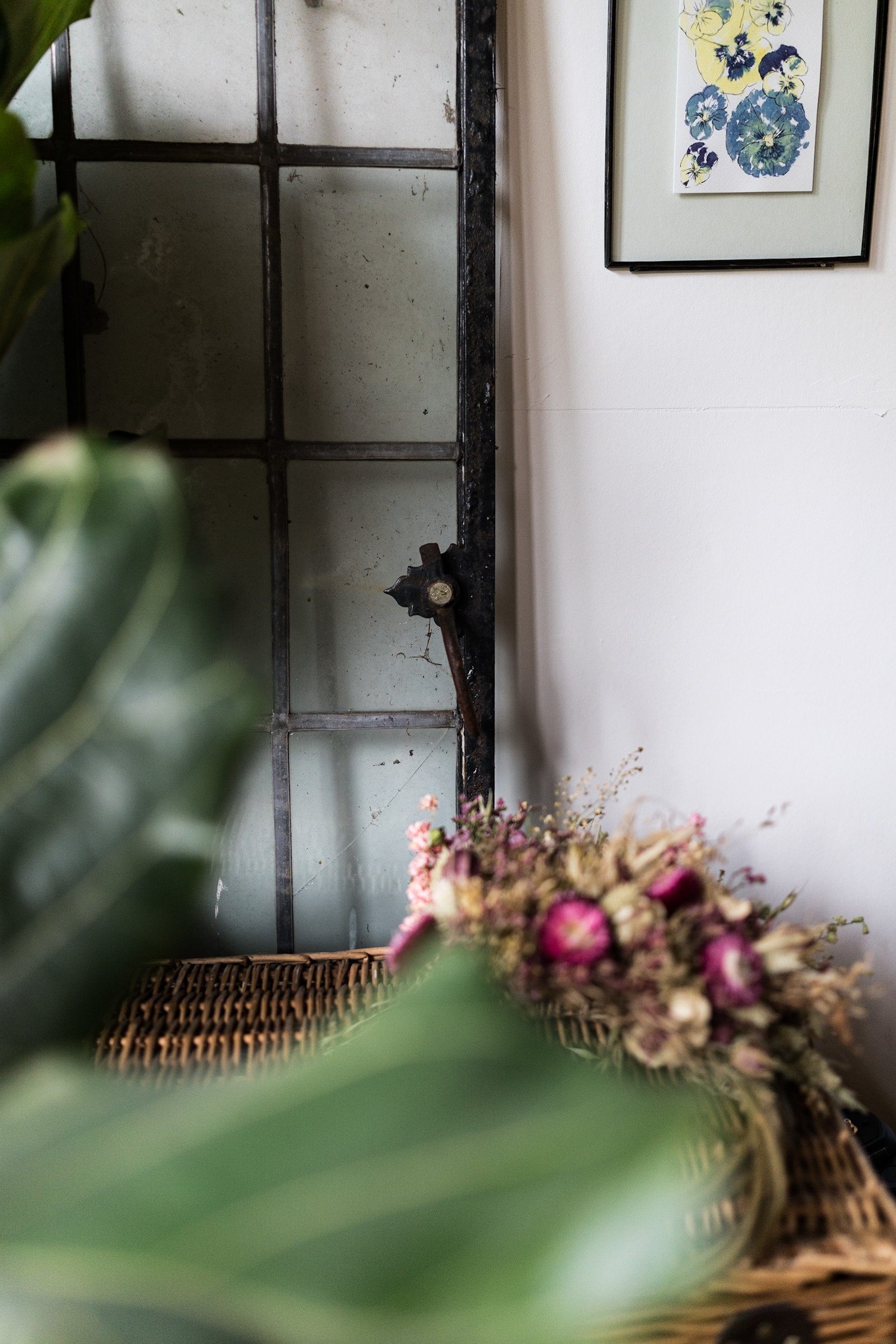 Interior of a room featuring an antique black iron window frame and a framed floral painting on a white wall, with a wicker basket and a bouquet of dried flowers in the foreground.