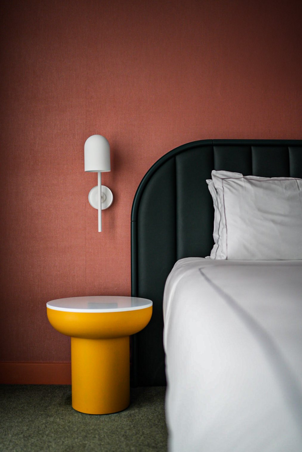 Close-up of a bedroom with a white pillow on a bed with white sheets, a green headboard, a yellow bedside table, and a white wall-mounted lamp against a pink textured wall.