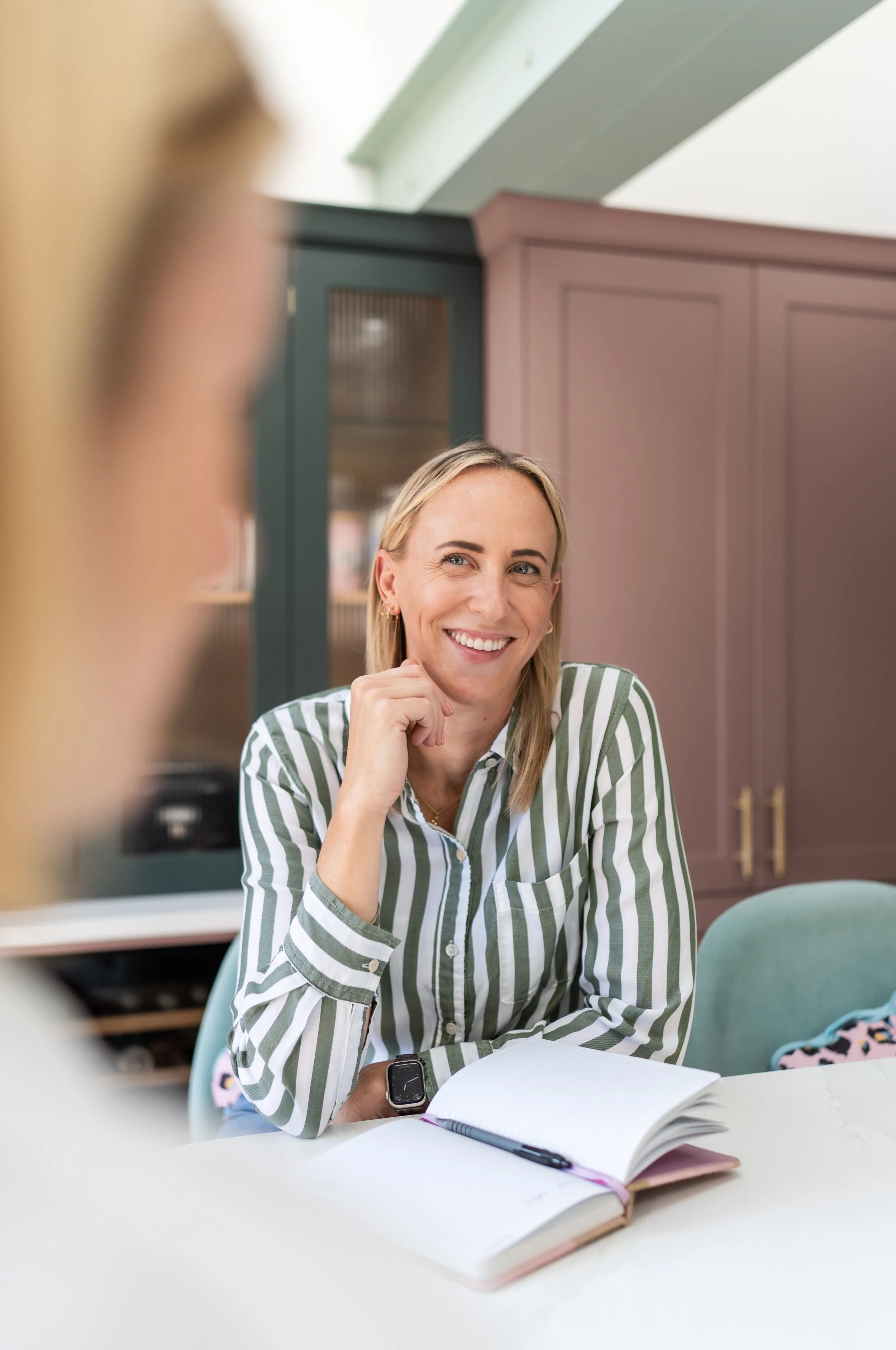 A woman with blonde hair, wearing a striped shirt, sitting at a table with an open notebook and pen, smiling and looking at someone off-camera indoors.