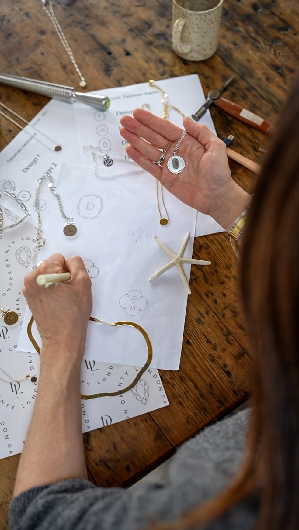 Person working on jewelry design with jewelry pieces, sketches, and tools on a wooden table.