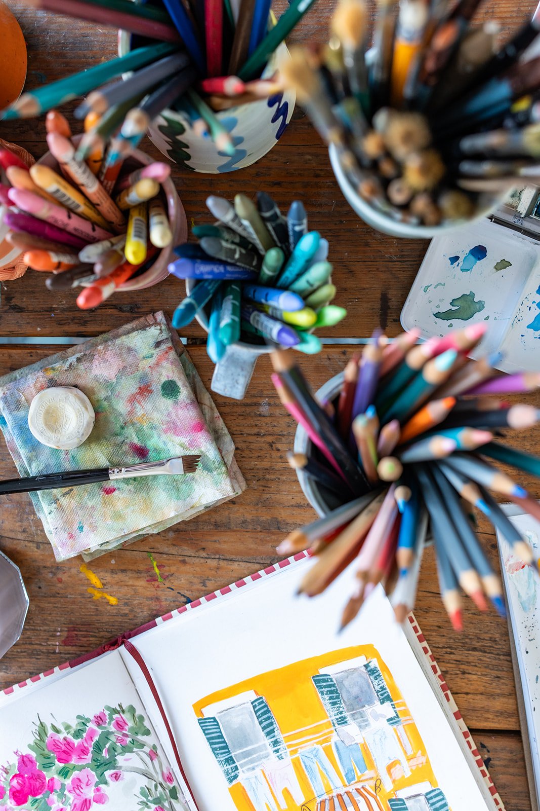 Overhead view of a wooden table with various art supplies, including colored pencils in cups, a paint palette, a paintbrush, a small container of white paint, and an open sketchbook with colorful illustrations.