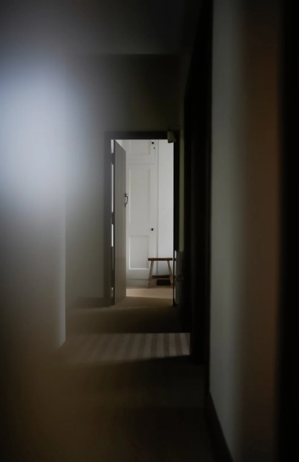 View down a dimly lit hallway with an open door at the end, revealing a room with a white door, a small wooden bench, and a piece of dark furniture.