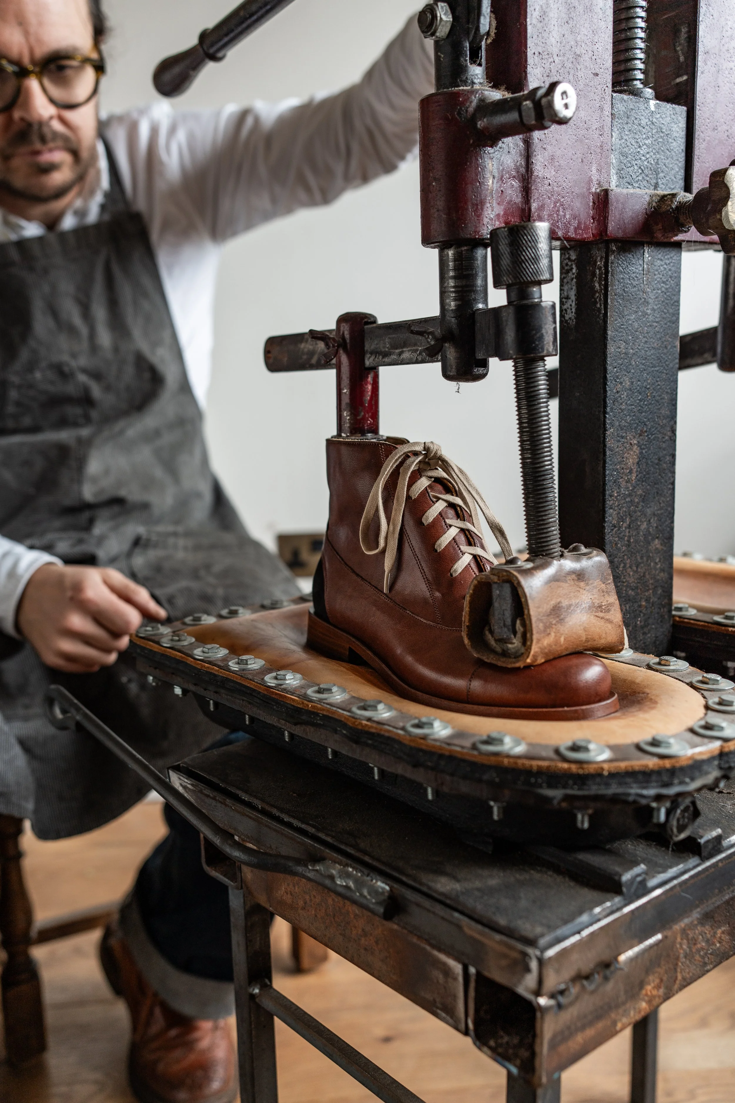 A person using a shoe-making machine to shape a brown leather boot with laces.