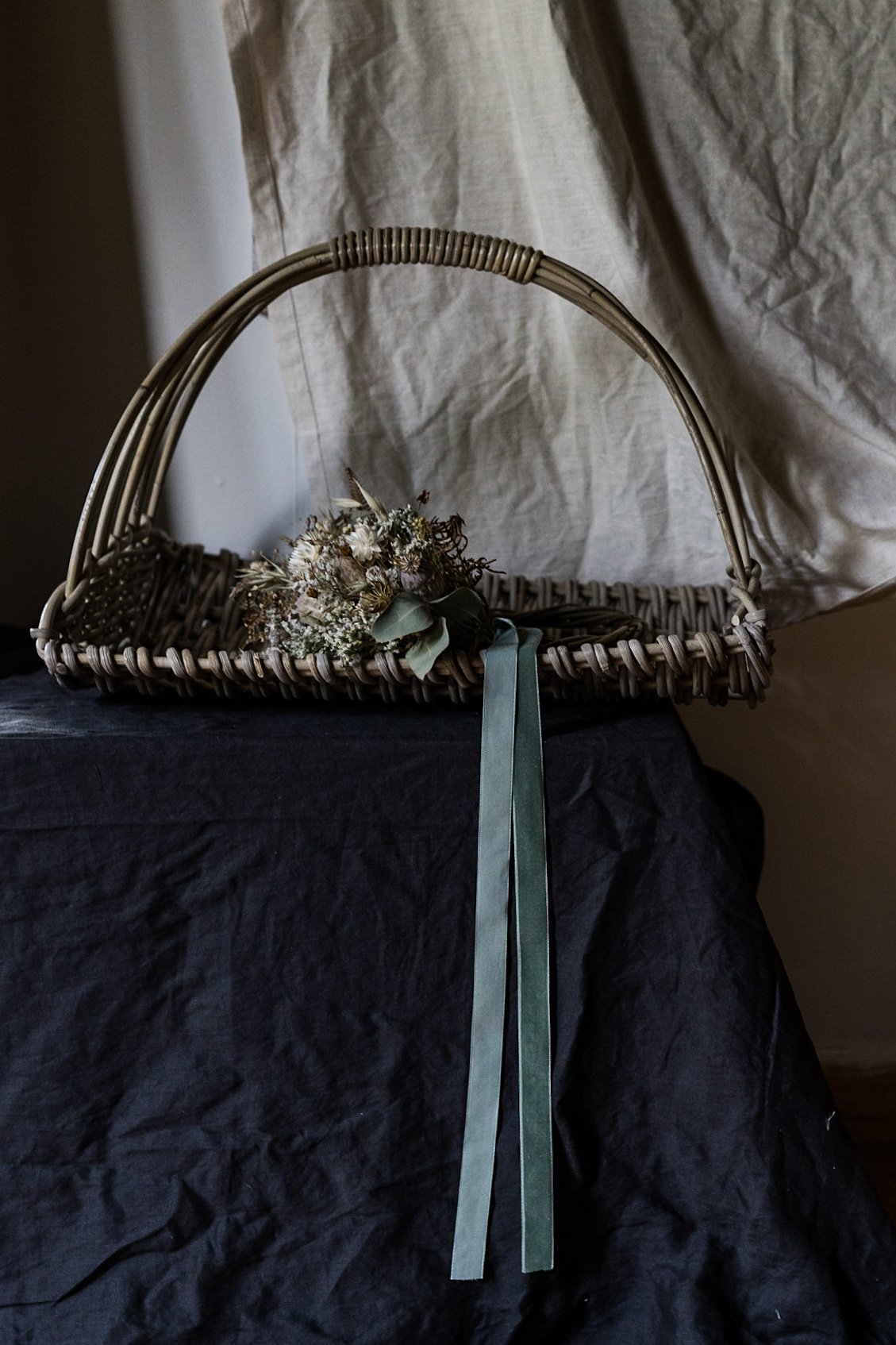 A wicker basket with a handle, decorated with dried flowers and green ribbons, placed on a black cloth-covered surface.