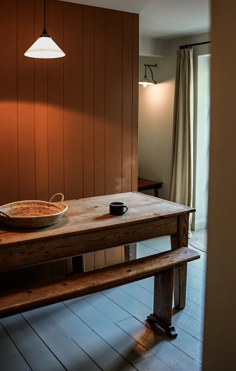 A rustic wooden dining table with a black mug and a woven basket inside a cozy dining area. The room has wood-paneled walls, a hanging pendant light, a window with curtains, and a small wall-mounted lamp.