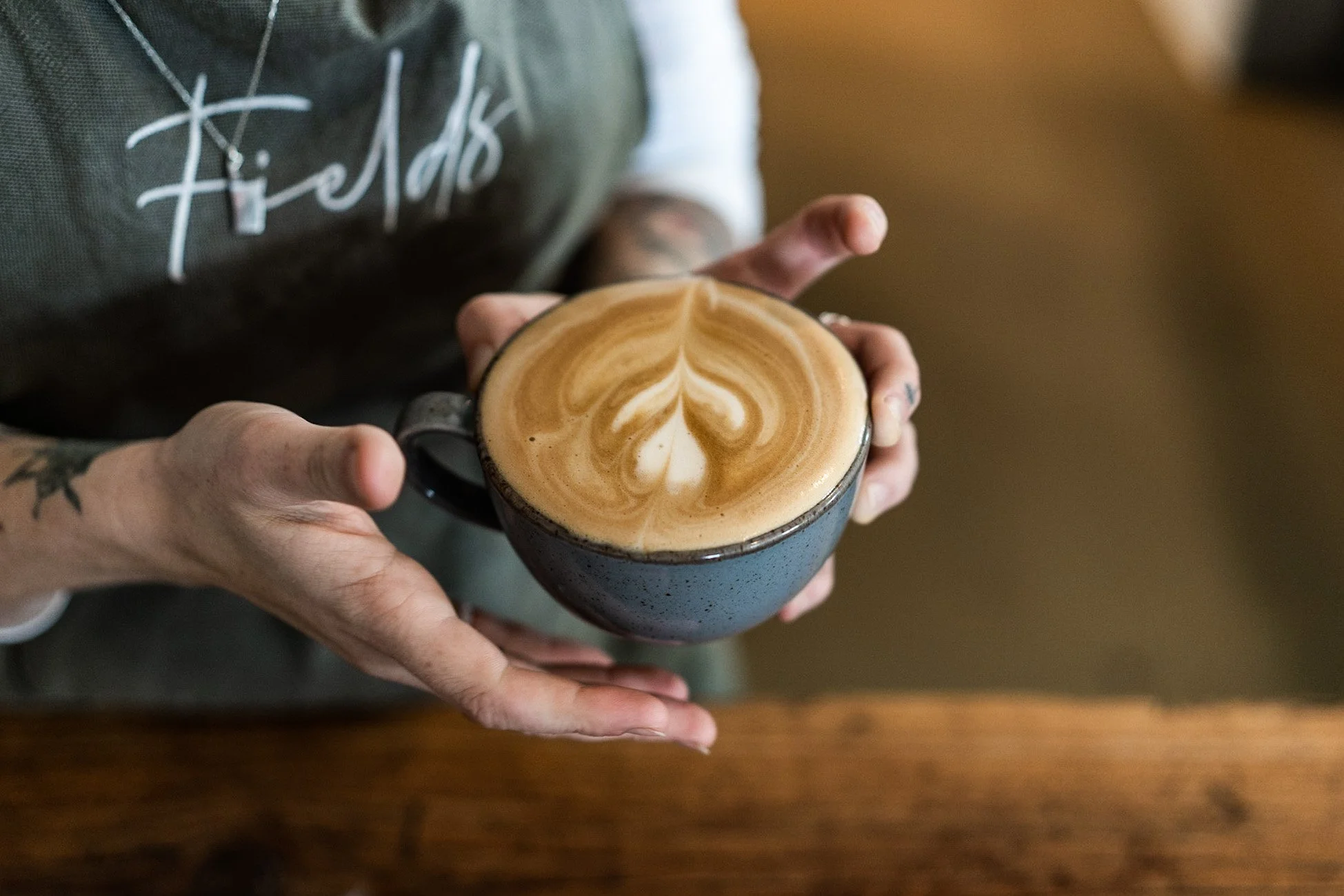 Person holding a black ceramic mug with a latte featuring latte art on top, in a cozy indoor setting.