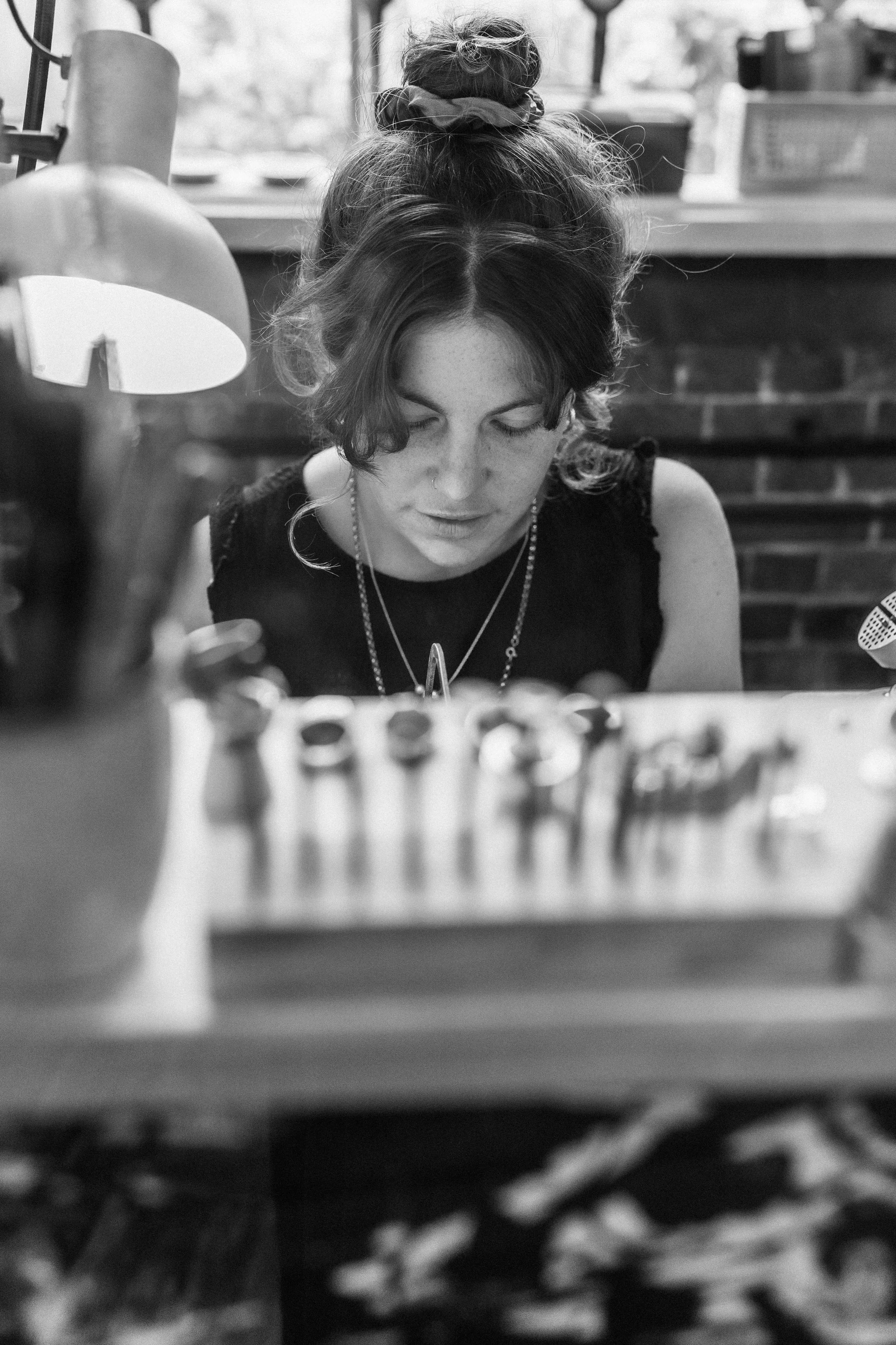 A woman with curly hair in a bun, wearing a black sleeveless top and layered necklaces, looking down at a work surface with jewelry tools and rings, in a workspace with brick walls.