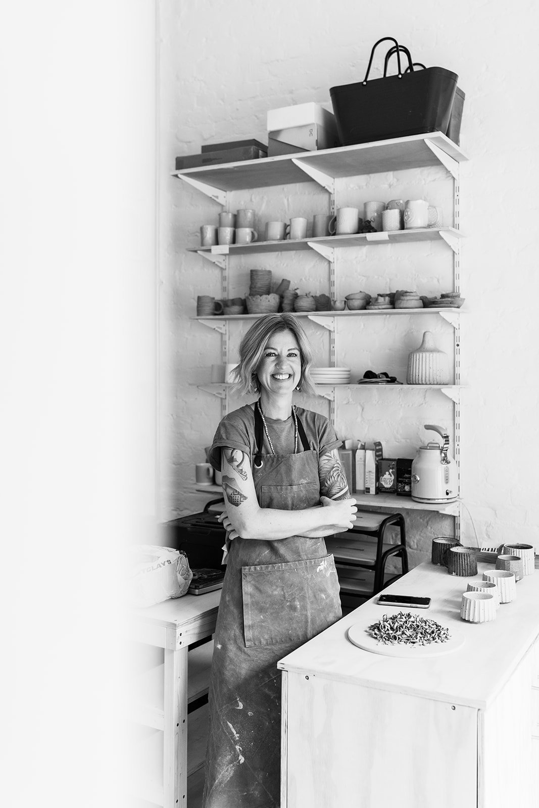 A smiling woman with tattoos wearing an apron, standing in a kitchen with shelves of pottery and mugs behind her.