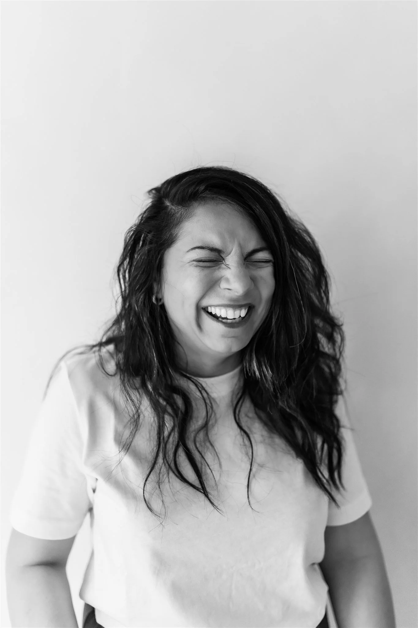 Black and white photo of a woman with long wavy hair laughing with her eyes closed, standing against a plain background.