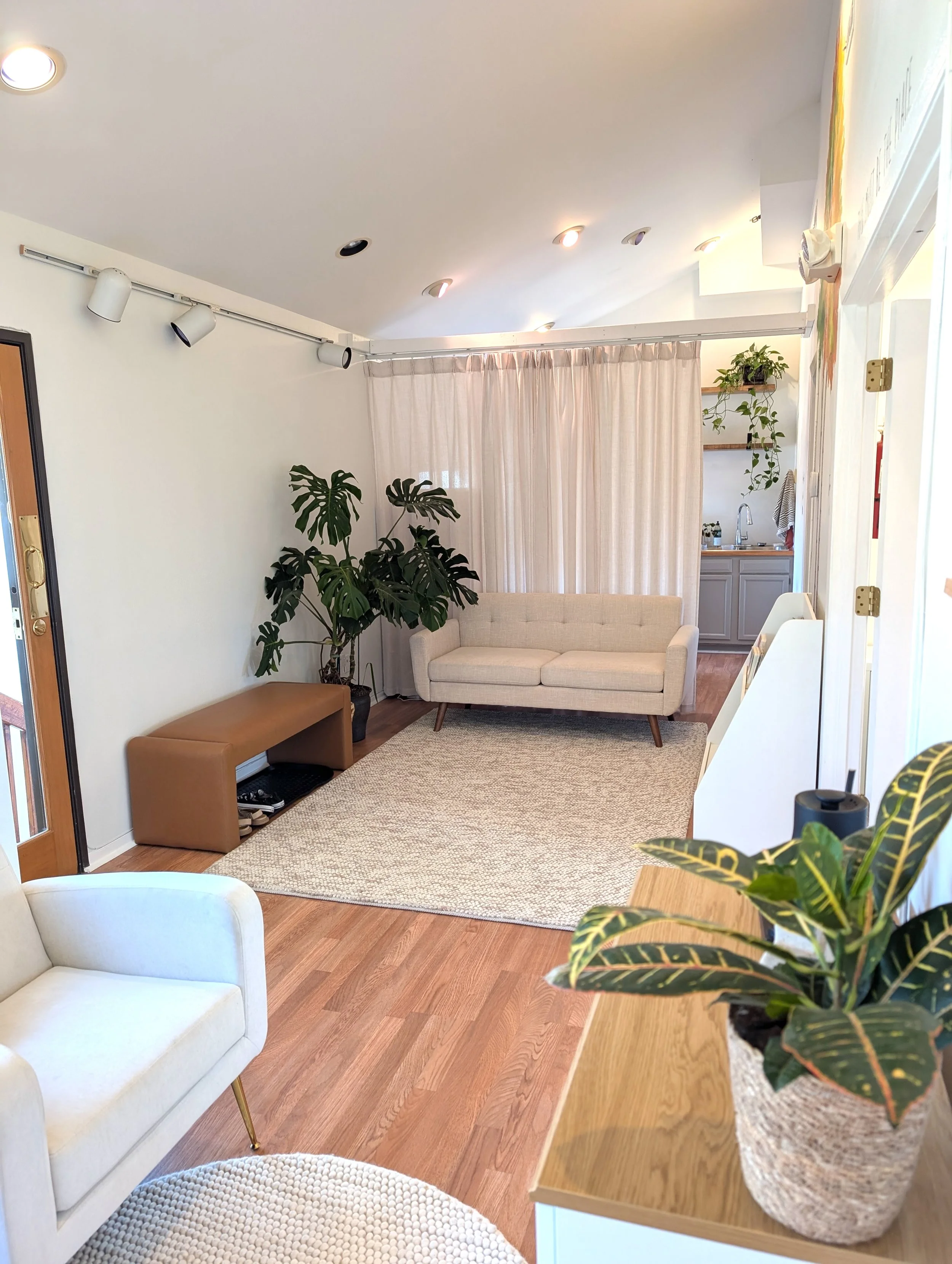A cozy living room area with a beige sofa, potted plants, a wooden console table, and a white armchair. The space has wooden floors, a woven rug, and is separated by a curtain from a small kitchenette area with shelves and plants. Recessed and track lighting illuminate the space.