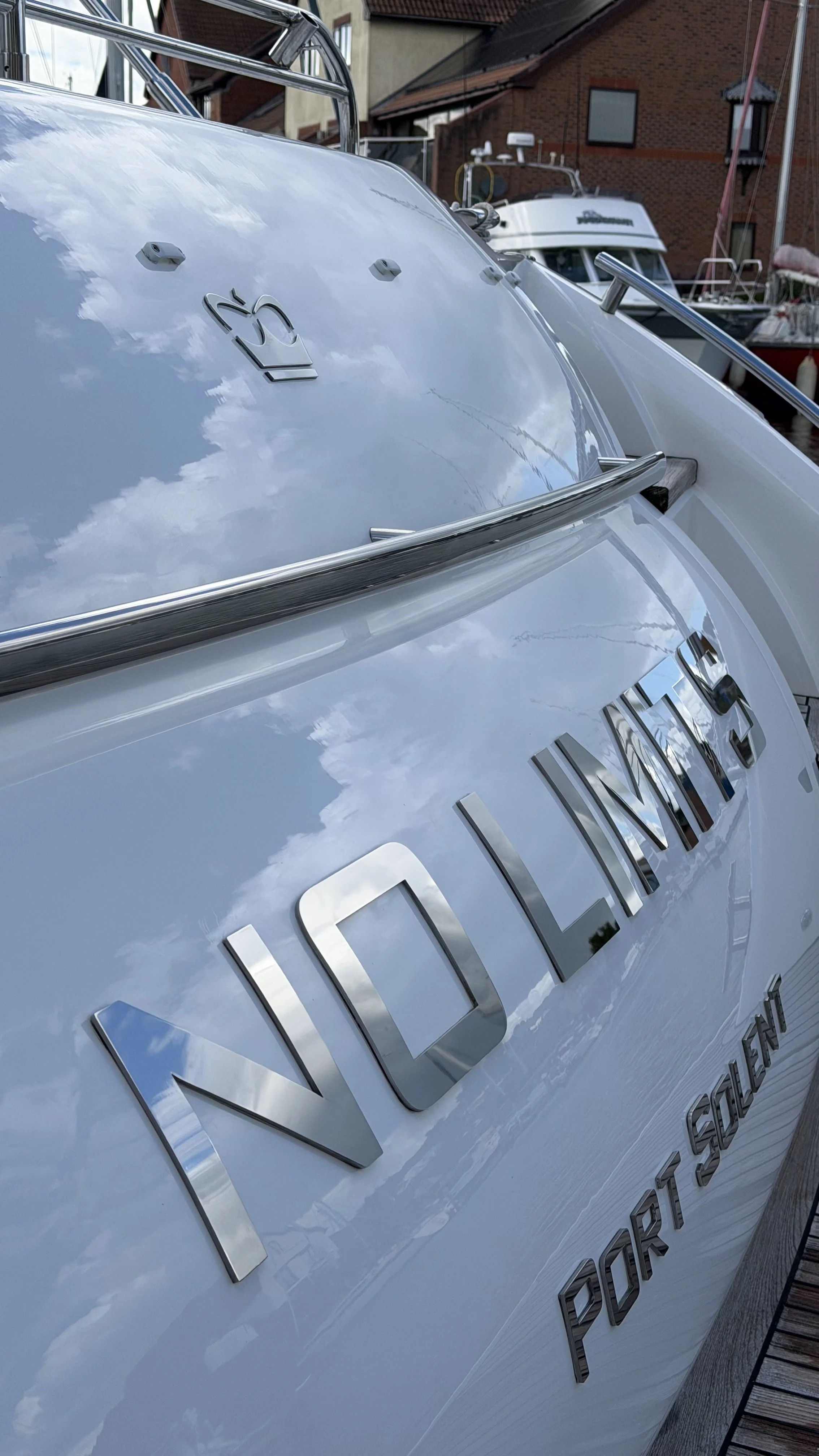 Close-up of a white boat with silver lettering and a logo, docked near a marina with other boats and brick buildings in the background.