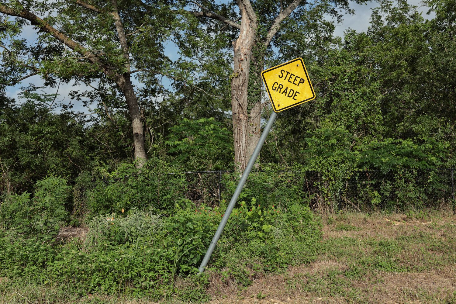 A yellow sign reading "steep grade" leans at an angle. Pecan trees in a Texas park are in the background.
