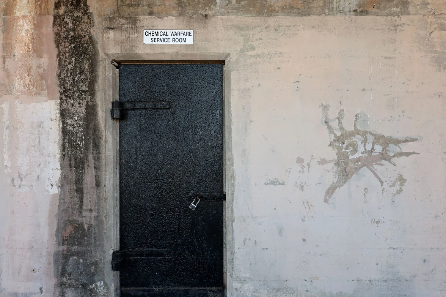 Image taken at Fort Travis in Texas showing a weathered concrete wall with a heavy locked iron door. There's a sign reading "chemical warfare service room" above the door.