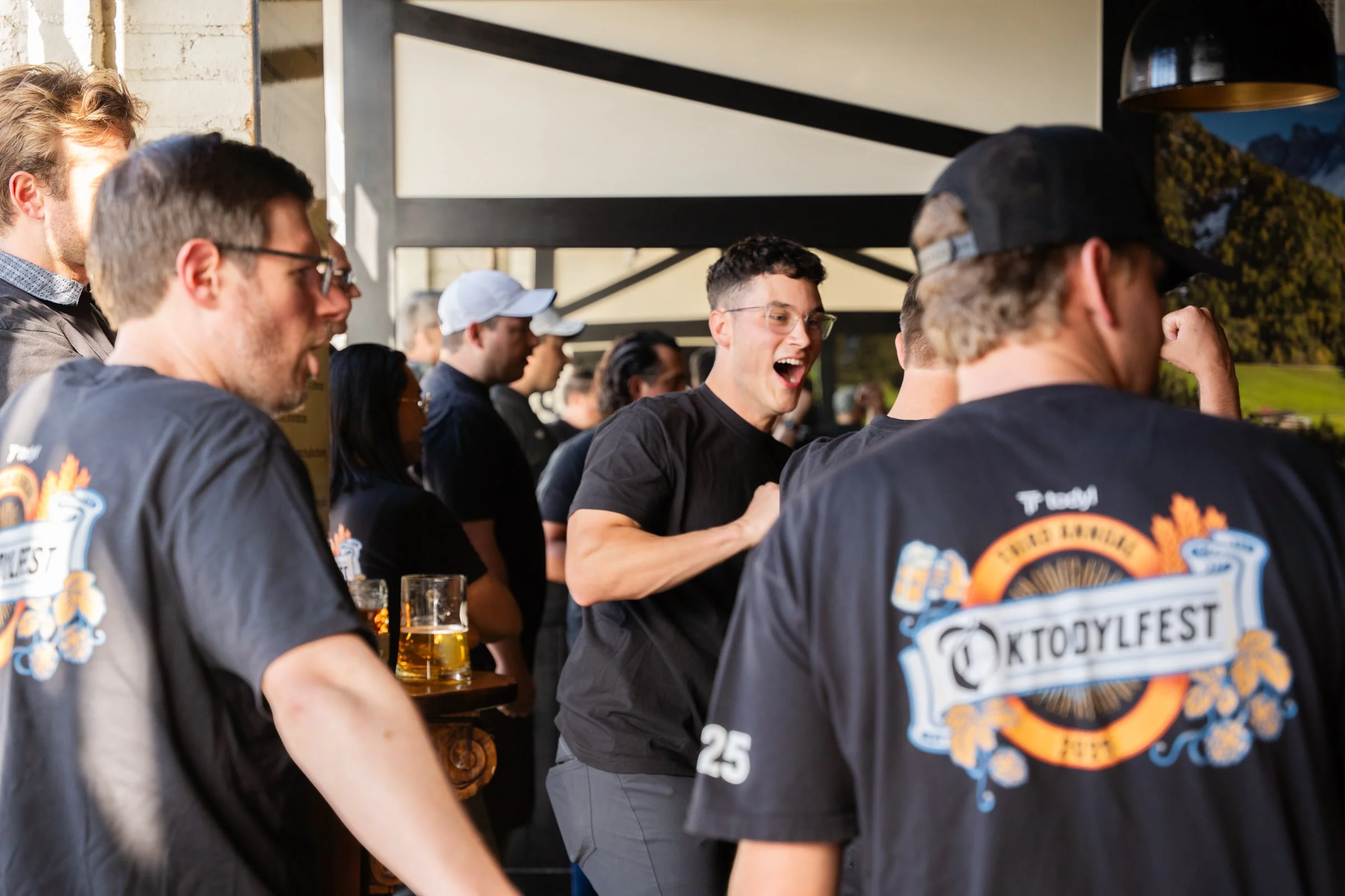 People gathered at an Oktoberfest beer festival, some wearing black t-shirts with Oktoberfest logos, one person is smiling and laughing, holding a drink, with others near tables and a scenic outdoor background.