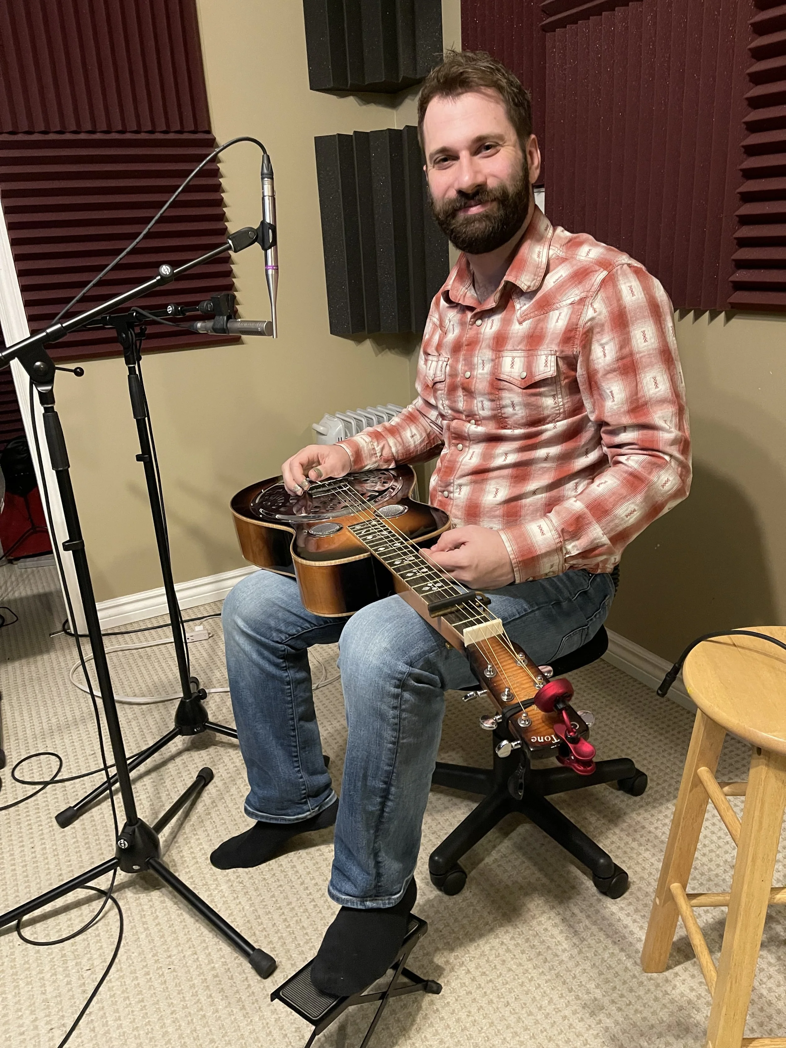 A man with a beard and mustache is sitting in a music studio, holding an acoustic guitar on his lap. He is wearing a red plaid shirt, jeans, and black socks, sitting on a chair with his feet on a pedal. There are acoustic foam panels on the wall behi