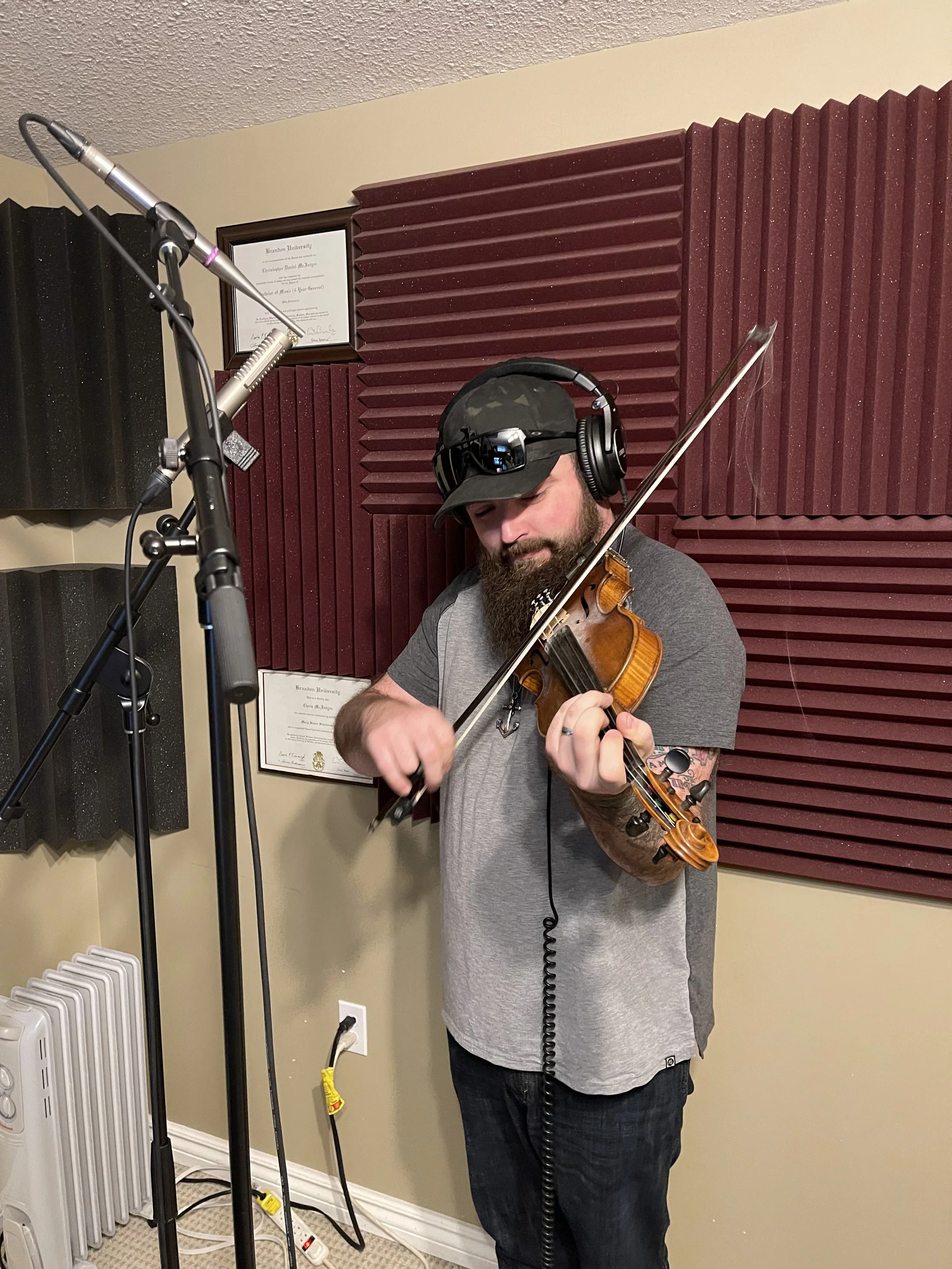 A man with a beard, wearing a black cap, headphones, and a gray t-shirt, is playing a violin in a recording studio with red and black soundproofing panels on the wall.