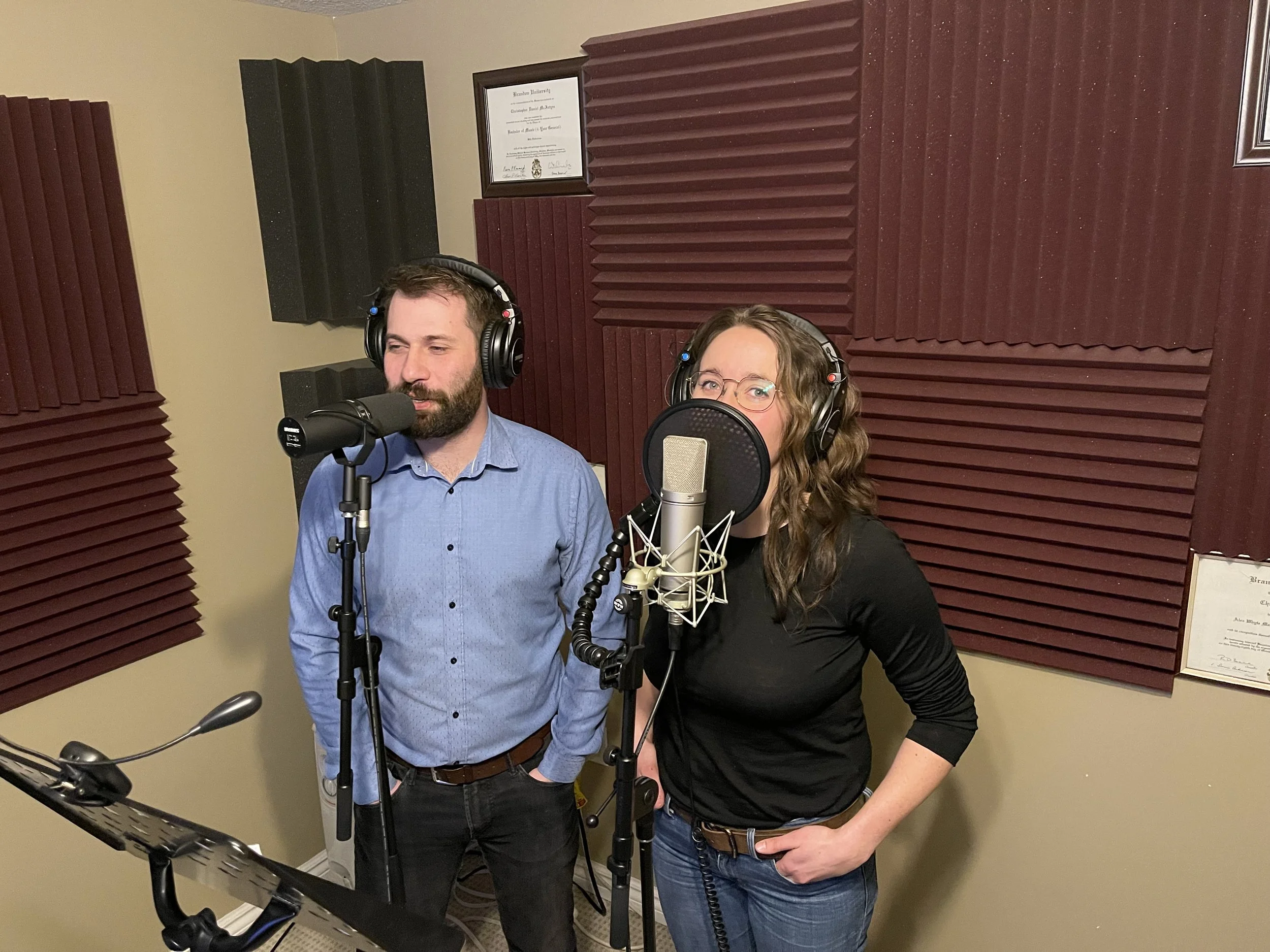 Two people, a man and a woman, are recording in a soundproof studio. They are standing in front of microphones with headphones on, and soundproof foam panels are on the walls around them.