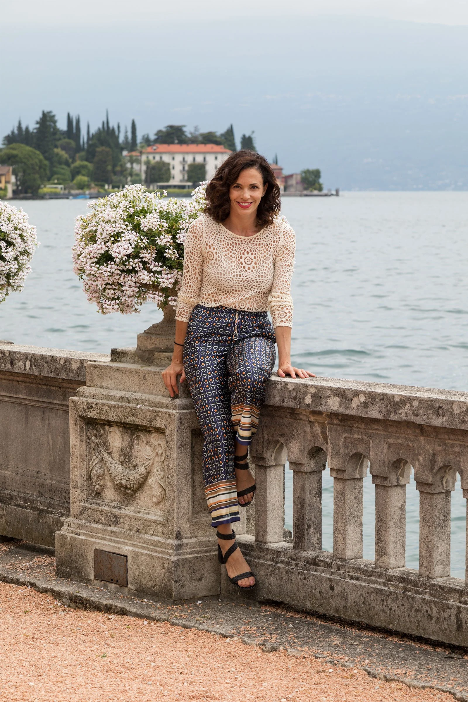 Smiling woman in patterned pants and a crochet top posing on a stone lakeside terrace with flowering shrubs and a villa in the background