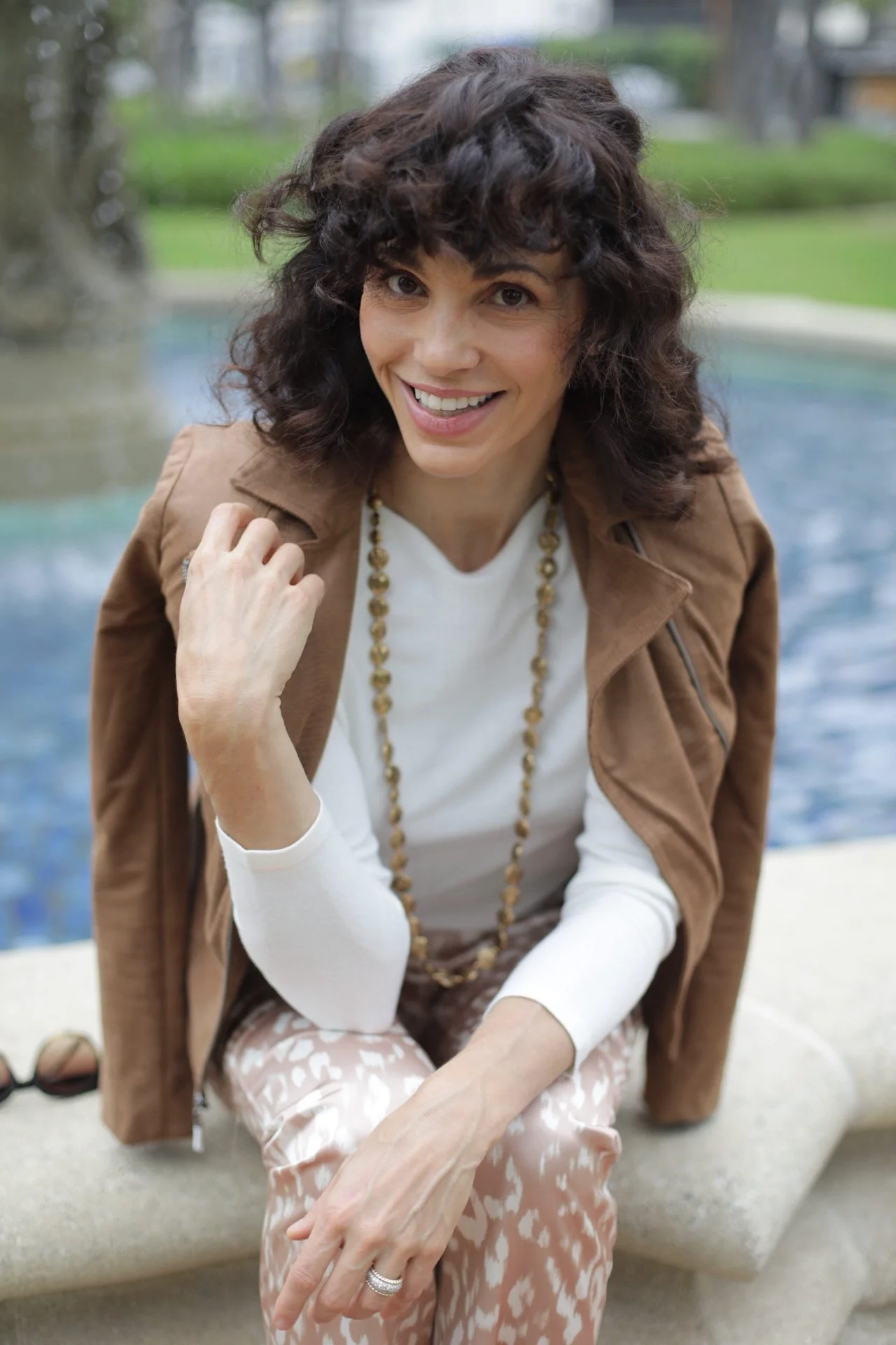 Smiling woman with curly dark hair wearing a brown jacket, white shirt, statement necklace, and patterned pants outdoors by a pool