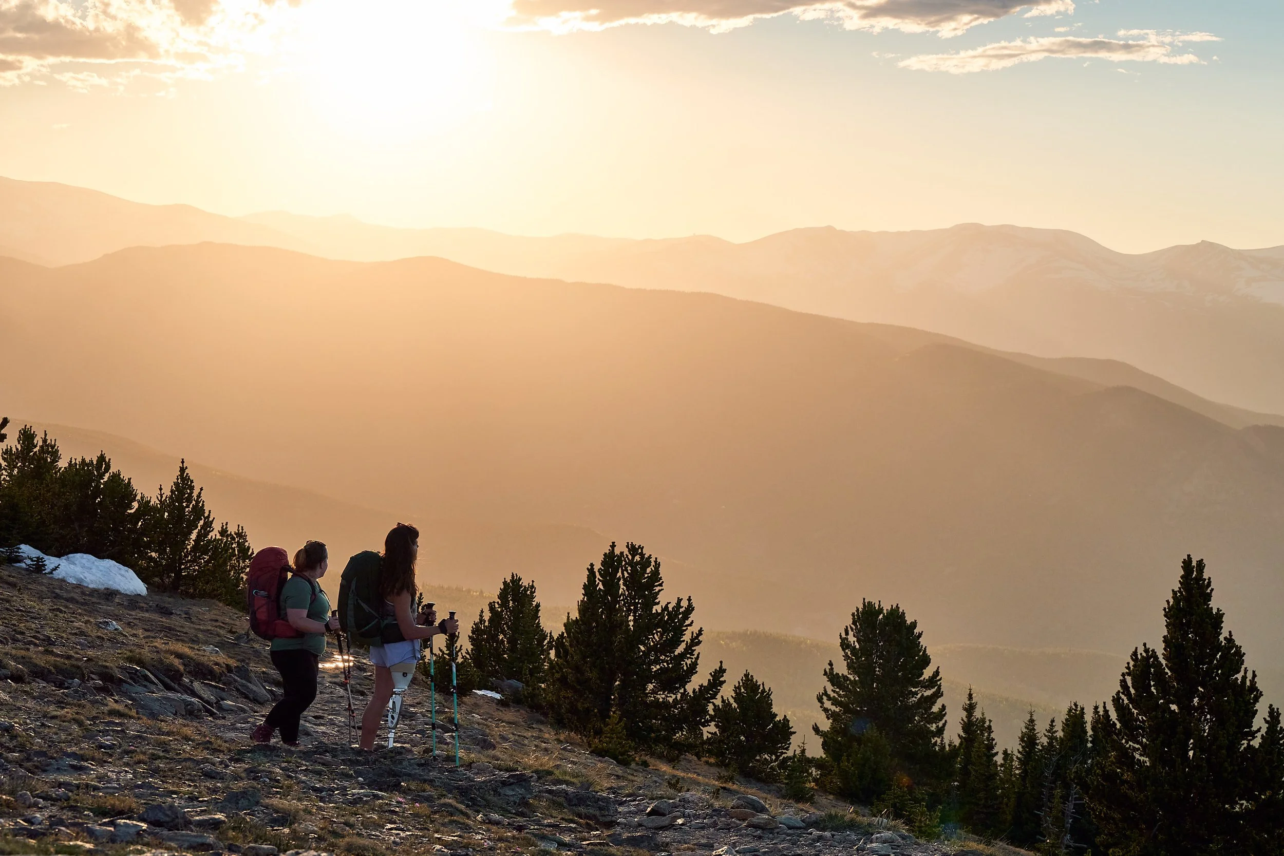 Two backpackers in Osprey packs pausing on an alpine slope at hazy sunset, outdoor lifestyle photography for Osprey Packs