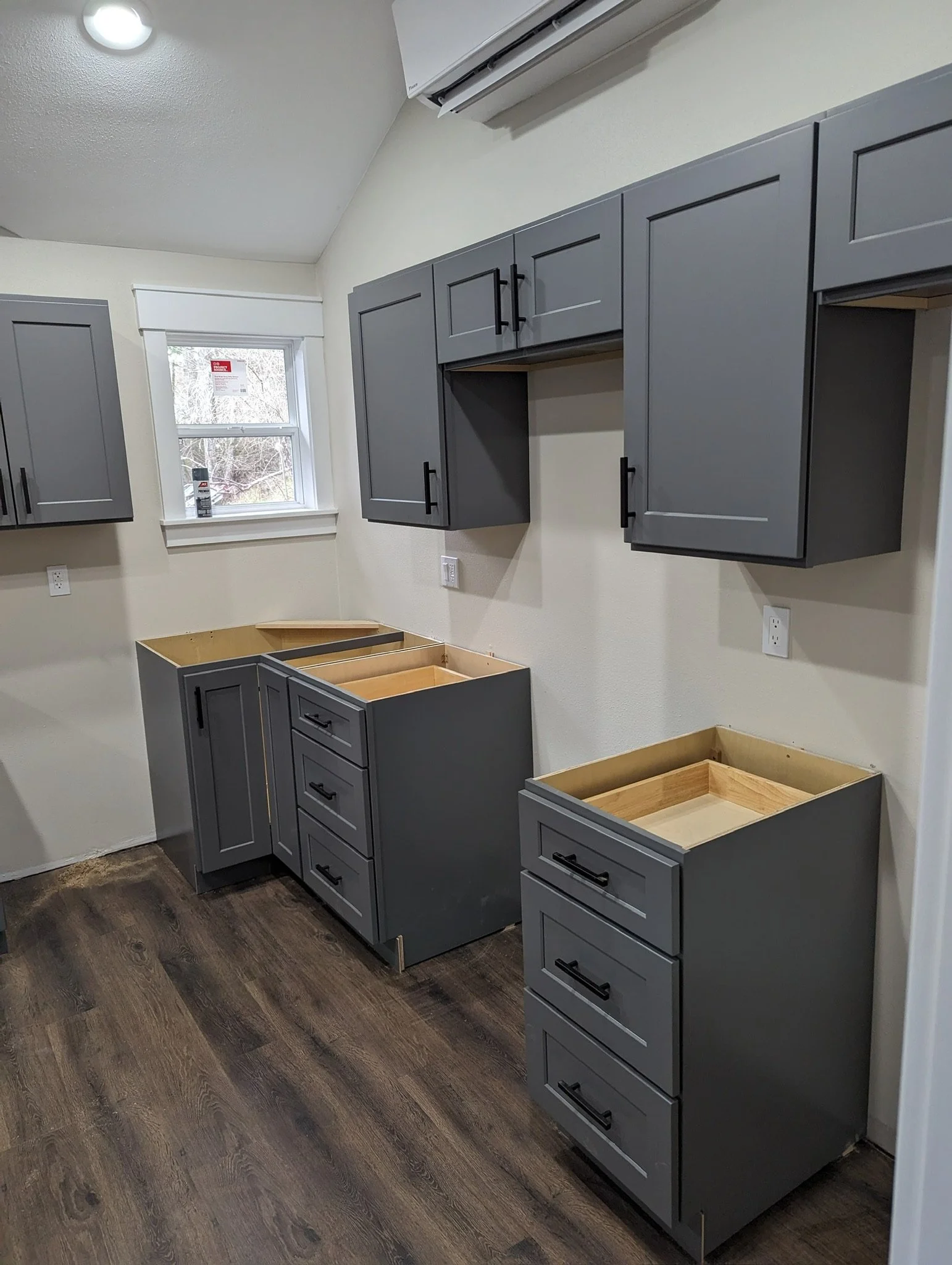 Kitchen cabinets installed in a kitchen under construction, with wall-mounted gray cabinets, base gray cabinets without countertops, wood flooring, a window, and an air conditioning unit above.