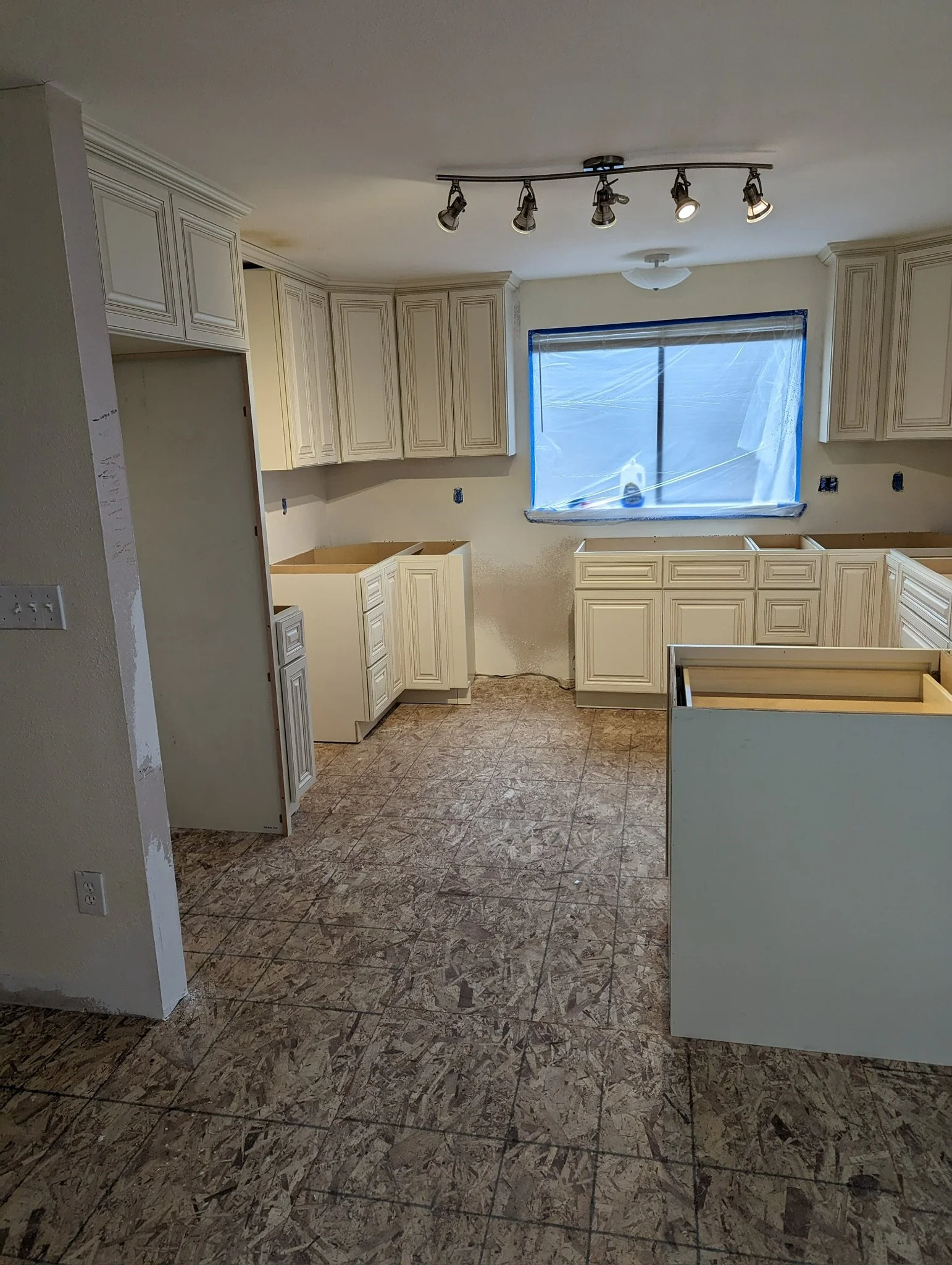 Kitchen under renovation with white cabinetry, a window covered with blue plastic, unfinished countertops, and plywood flooring.
