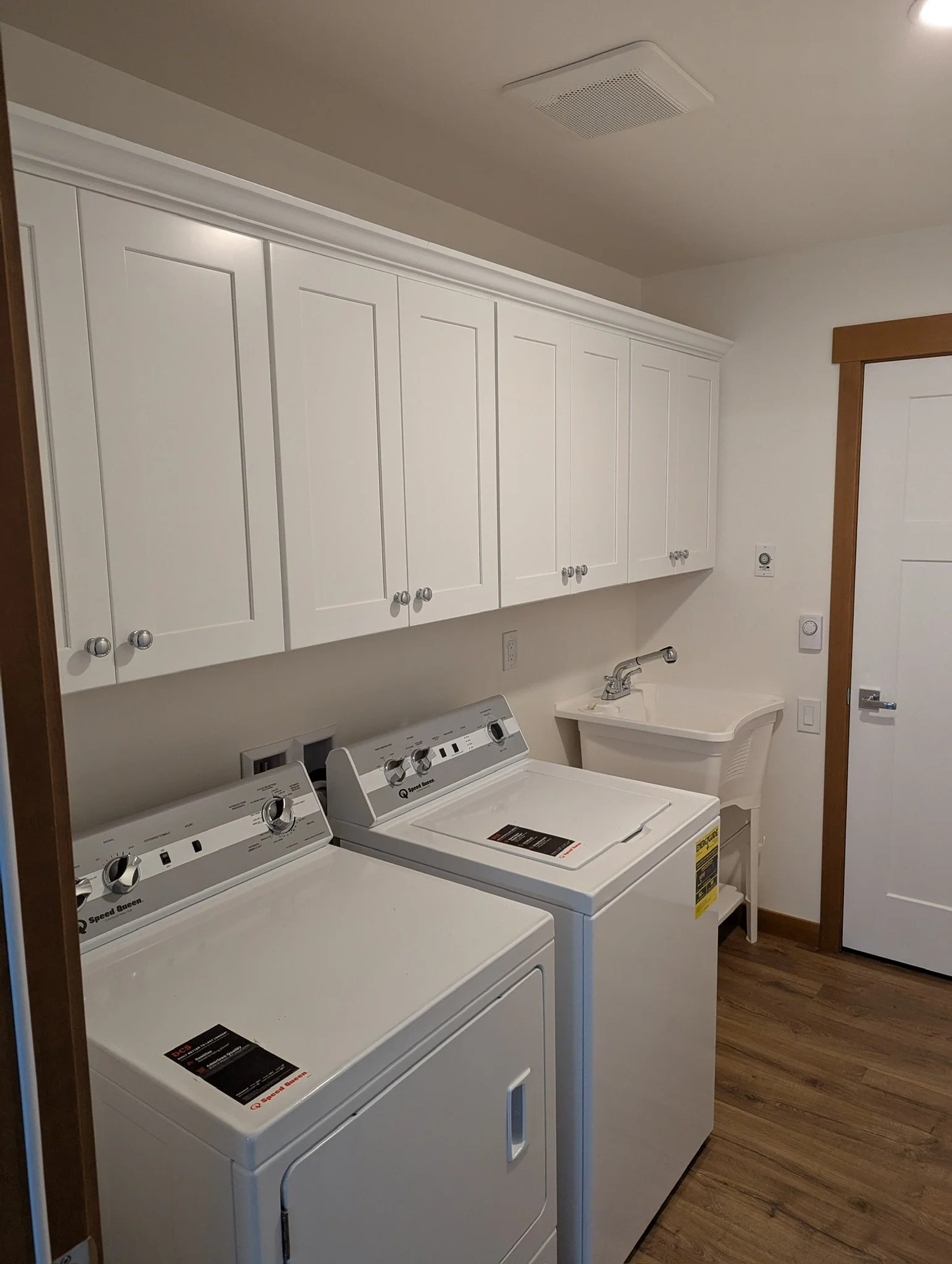 A laundry room with white washing machine and dryer, white cabinets above, a utility sink, and wood flooring.