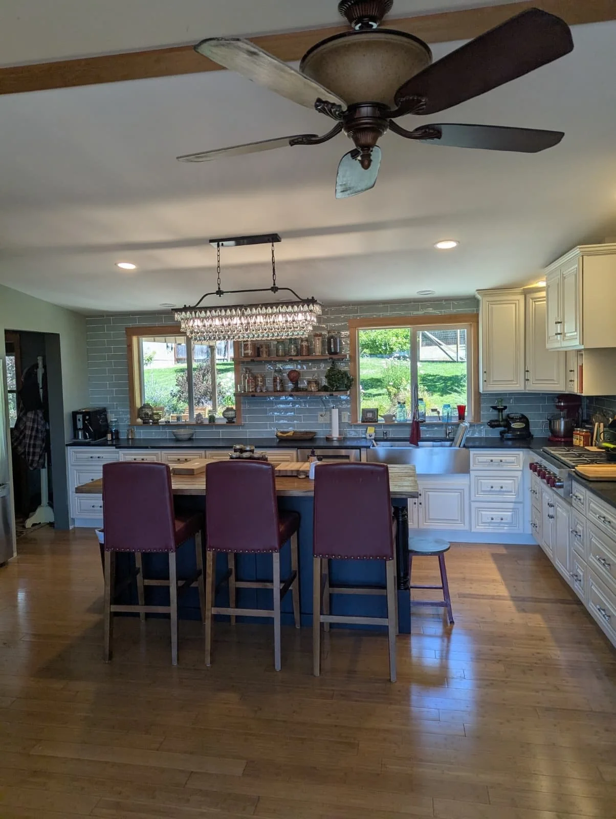 Bright kitchen with white cabinets, a wooden island with seating, a chandelier, and large windows showing a green outdoor area.