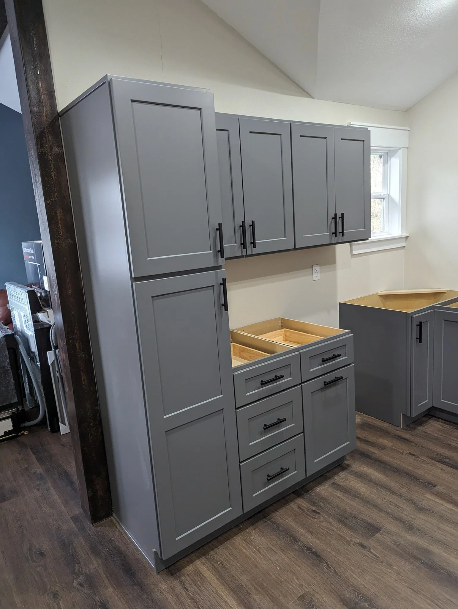 Kitchen cabinets with a gray finish and black handles, some drawers and cabinets are not fully installed, in a room with wood flooring and a window.