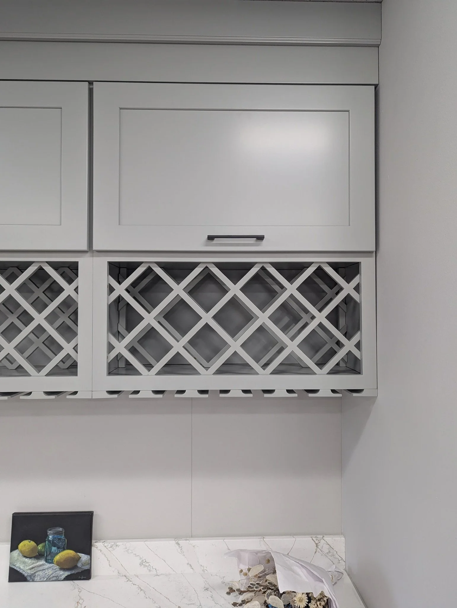 White kitchen cabinet with a black handle, a countertop with a framed picture of lemons and a jar, and a paper bag with seashells on it.