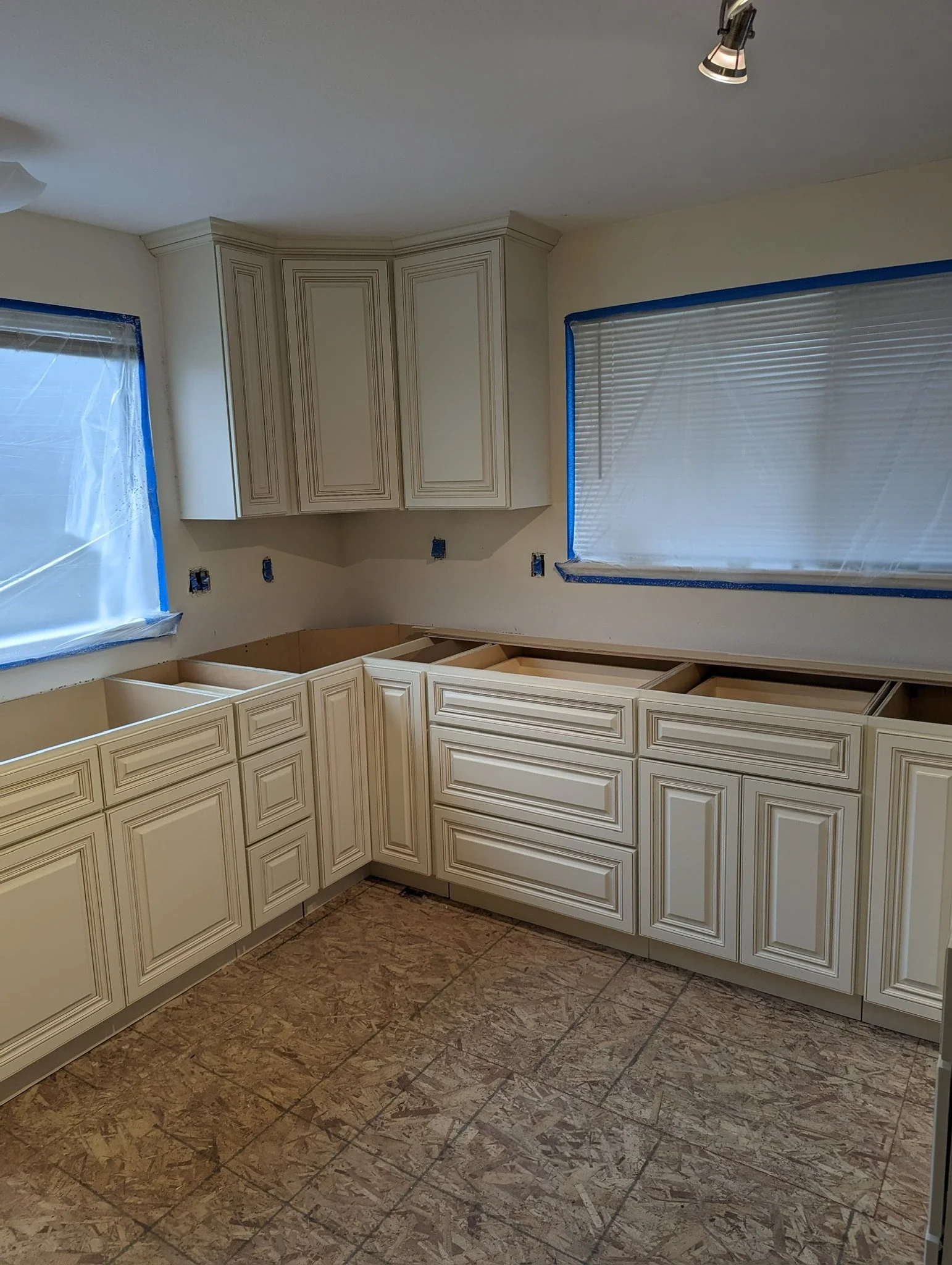 Kitchen under renovation with cream-colored cabinets, some upper cabinets installed, and walls protected with plastic coverings on windows.